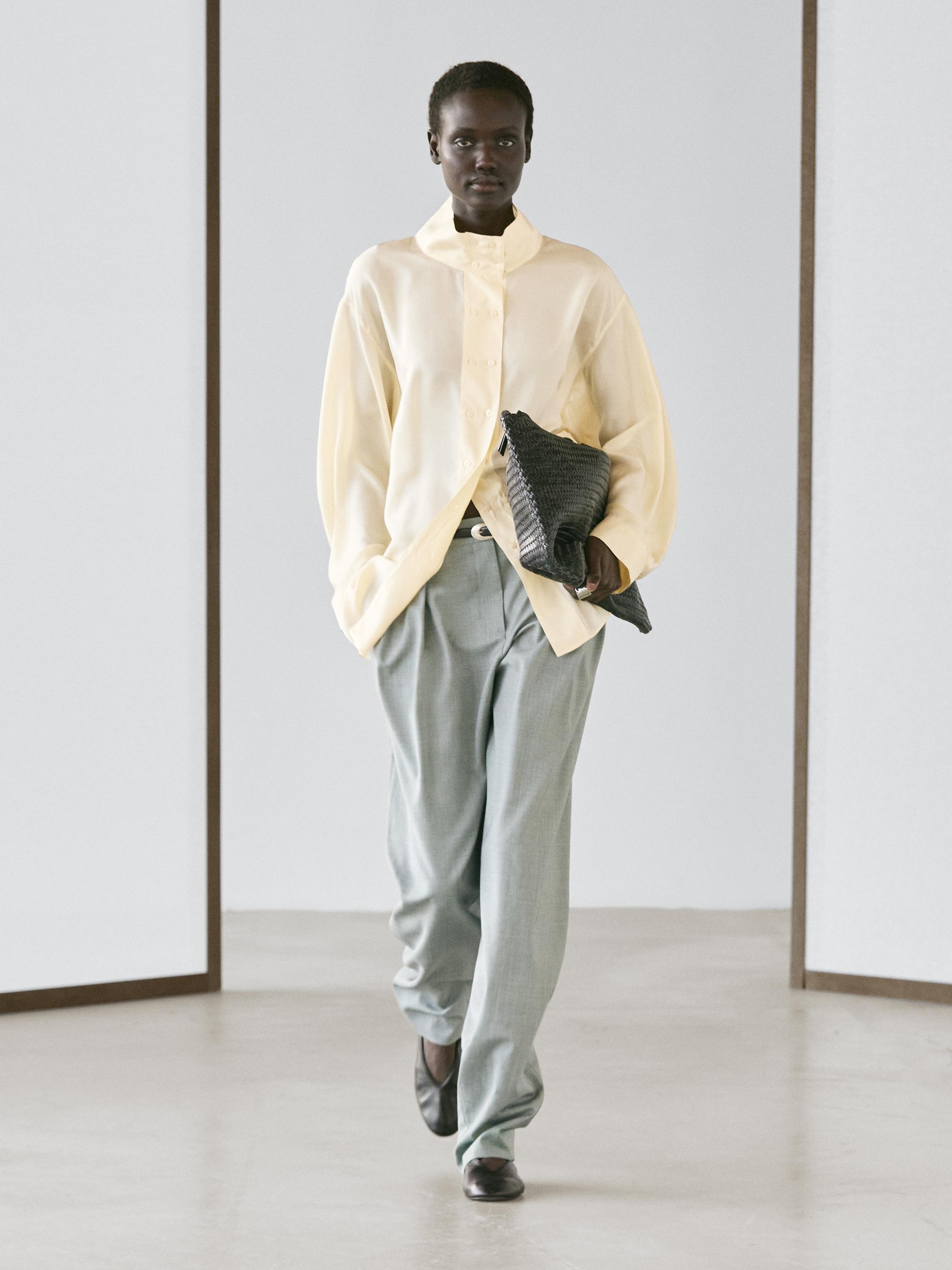 Ensemble composé d'une chemise boutonnée beige et d'un pantalon fluide gris-bleu à pinces, poches latérales et chaussures noires.