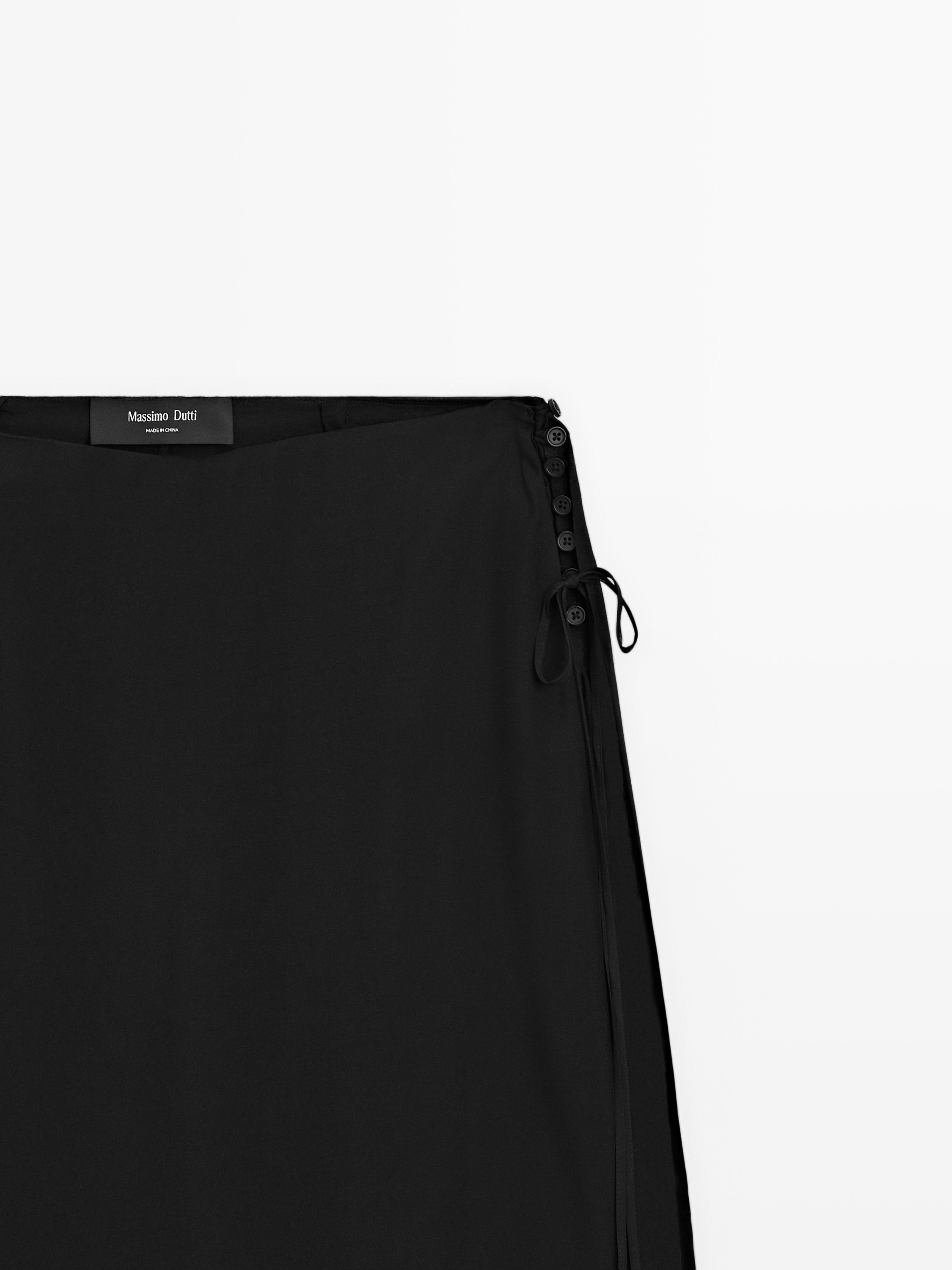 Λεπτομέρεια του πάνω μέρους μιας μαύρης φούστας με πλαϊνά κουμπιά και φιόγκο.