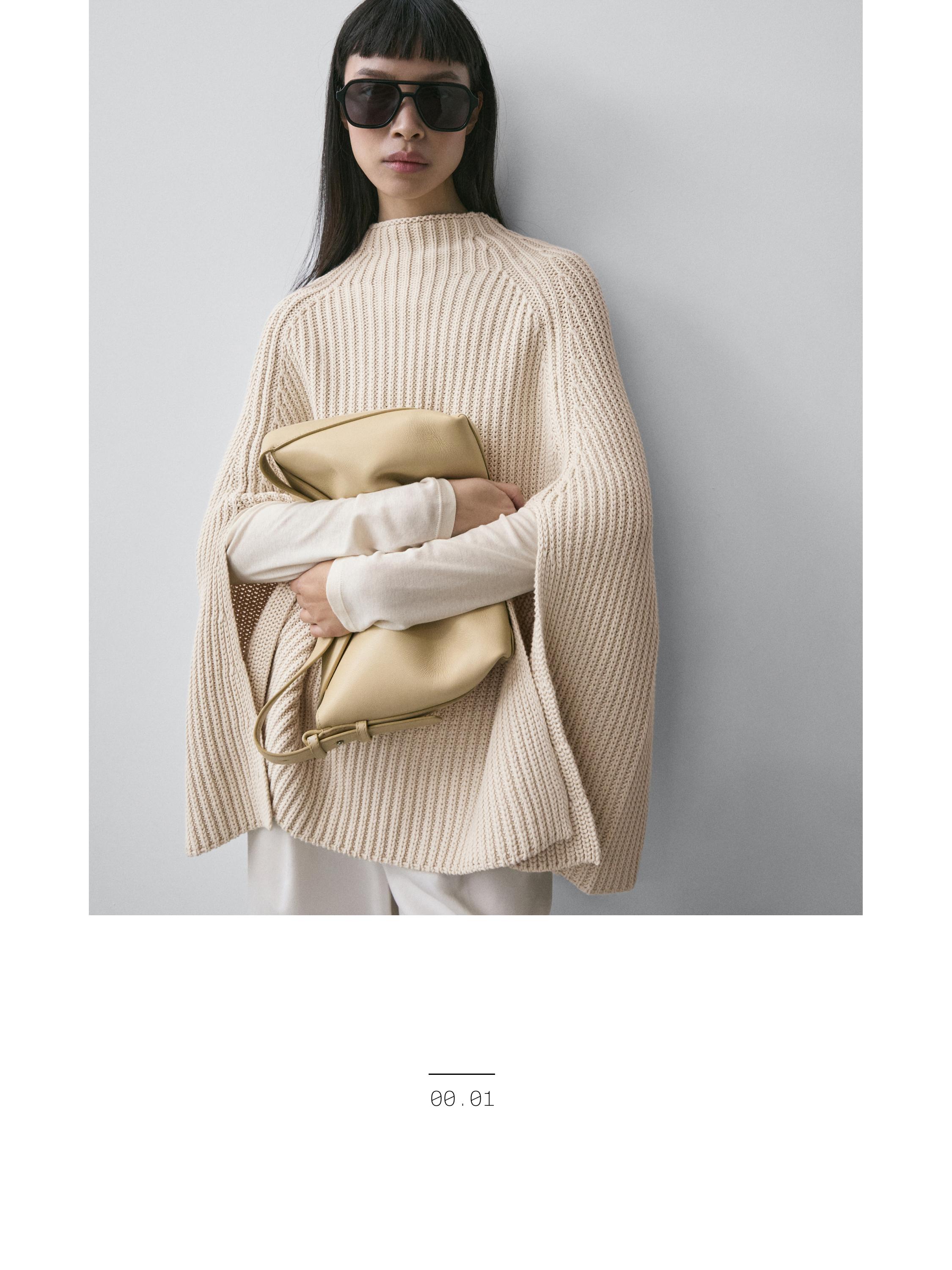 طقم مكون من كاب بيج محبوك سميك وبنطلون أبيض. تحمل حقيبة يد كبيرة بيج ونظارات شمسية داكنة.