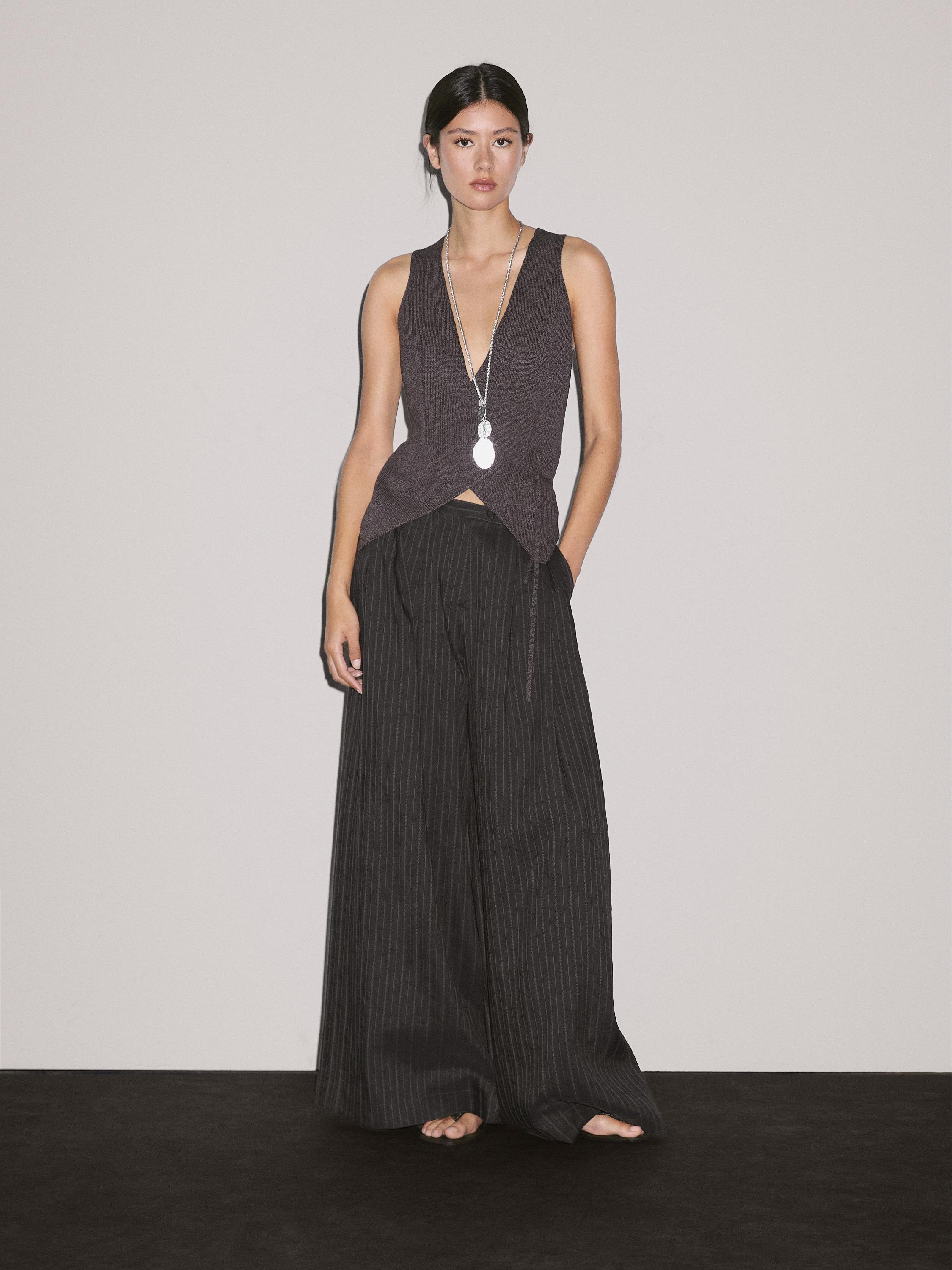 Setelan rompi silang tanpa lengan warna vanila dan celana hitam model lebar dengan garis-garis vertikal tipis.