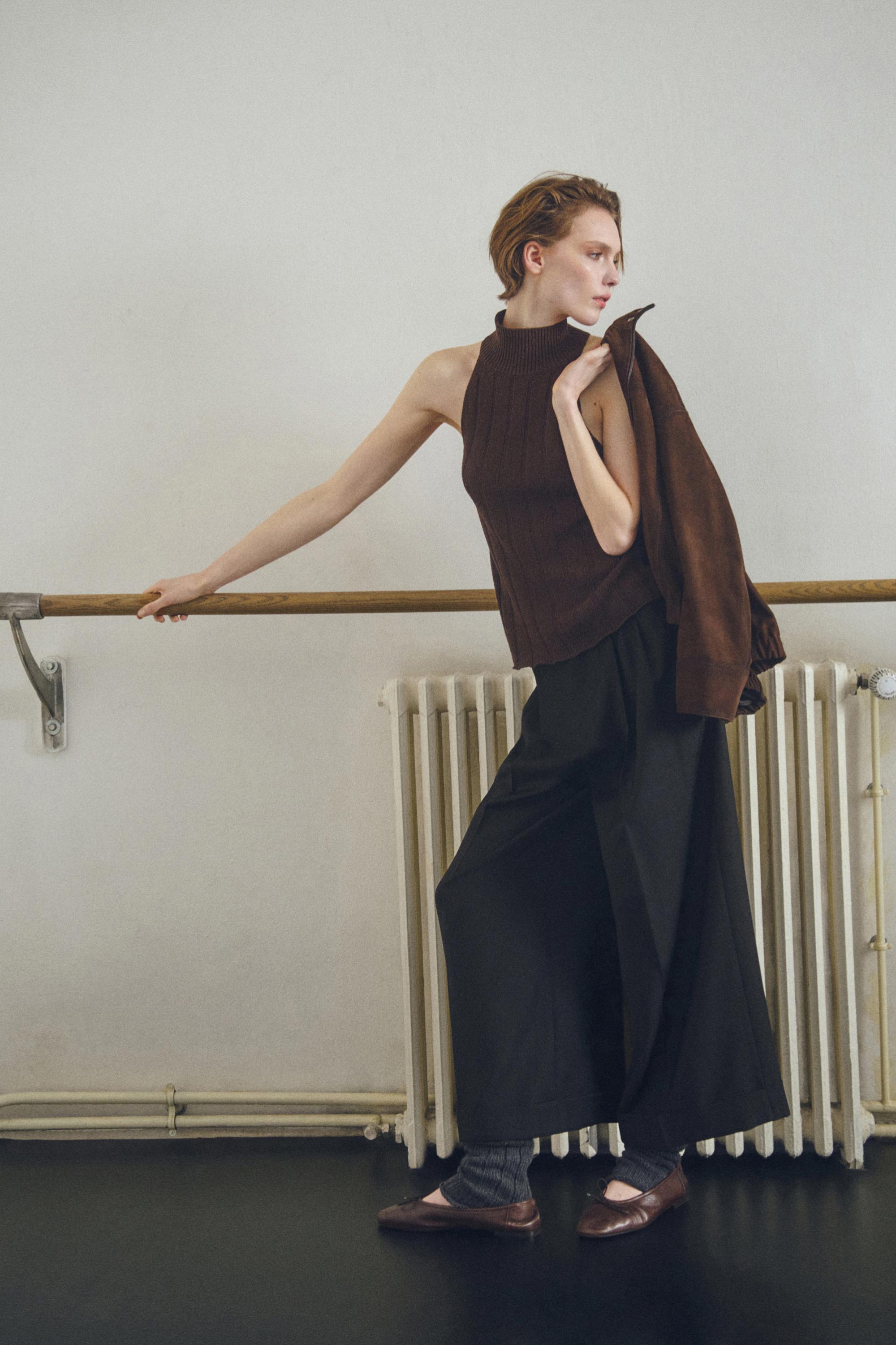 Set aus ärmellosem, schokoladenbraunem Neckholder-Top und schwarzer weiter Hose. Trägt eine braune Jacke über der Schulter.