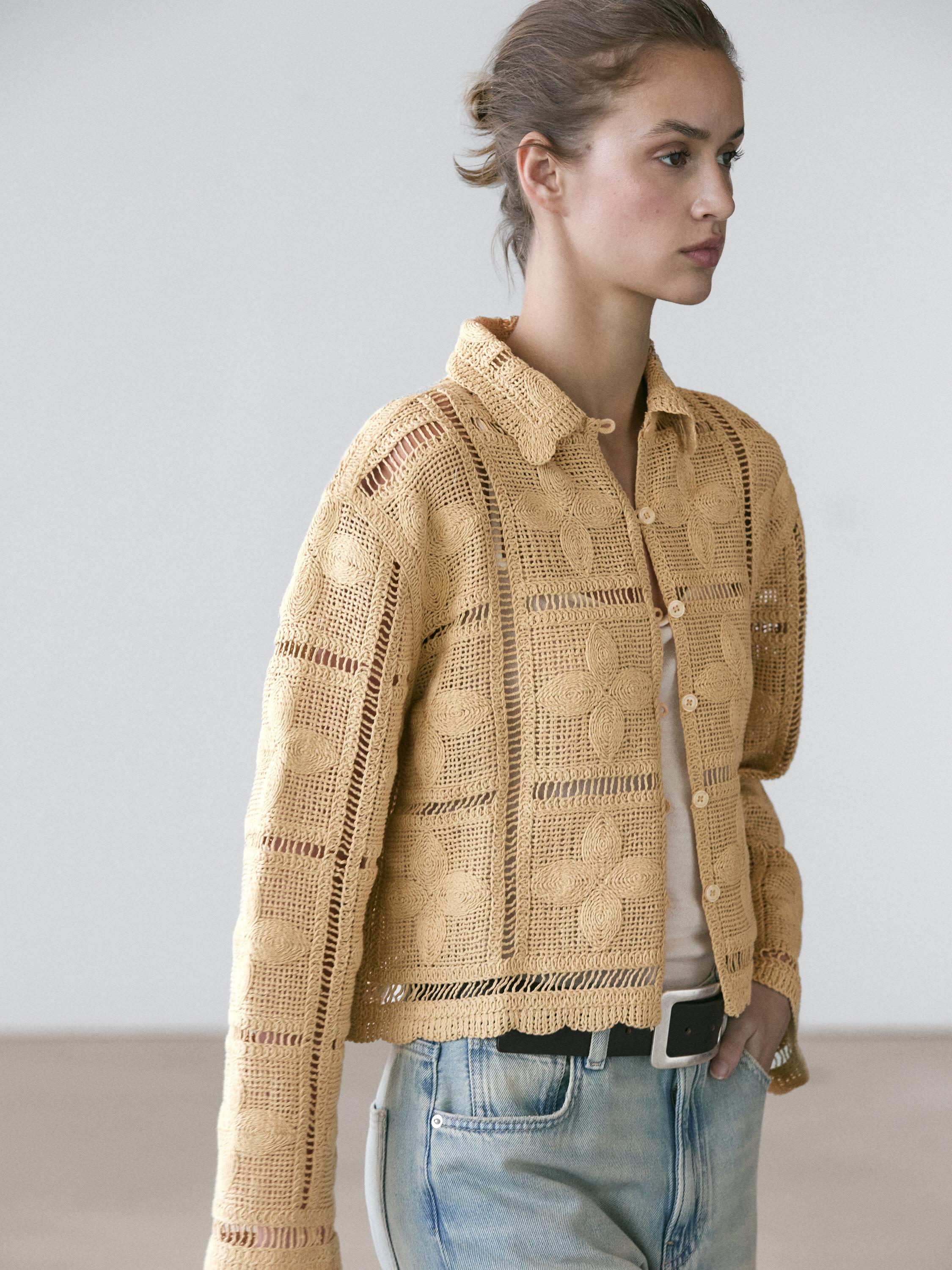 Halftotaal van een vanillekleurig gehaakt jasje met knopen en een opengewerkt bloemmotief, over een wit T-shirt en jeans met zijzak.