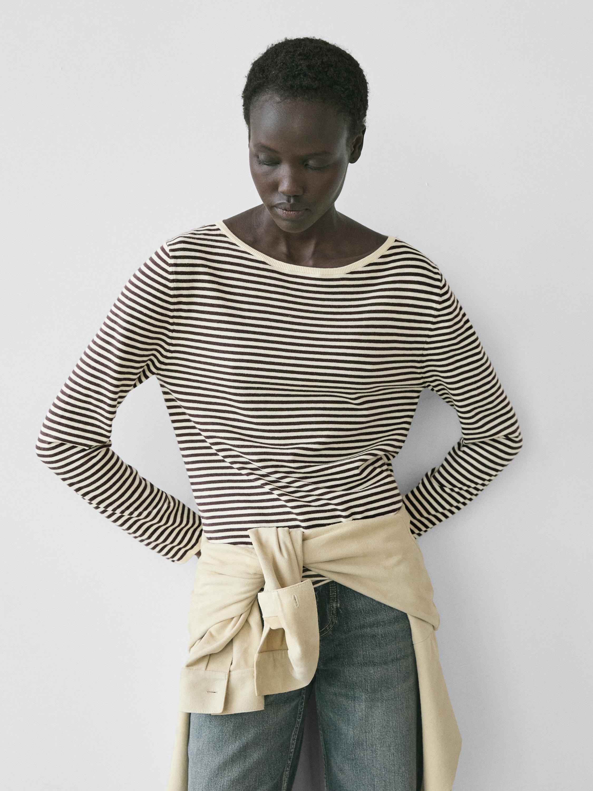 Langärmeliges, gestreiftes Oberteil in Ecru und Schwarz, mit blauen Jeans und einem beigen Sweatshirt um die Taille gebunden.