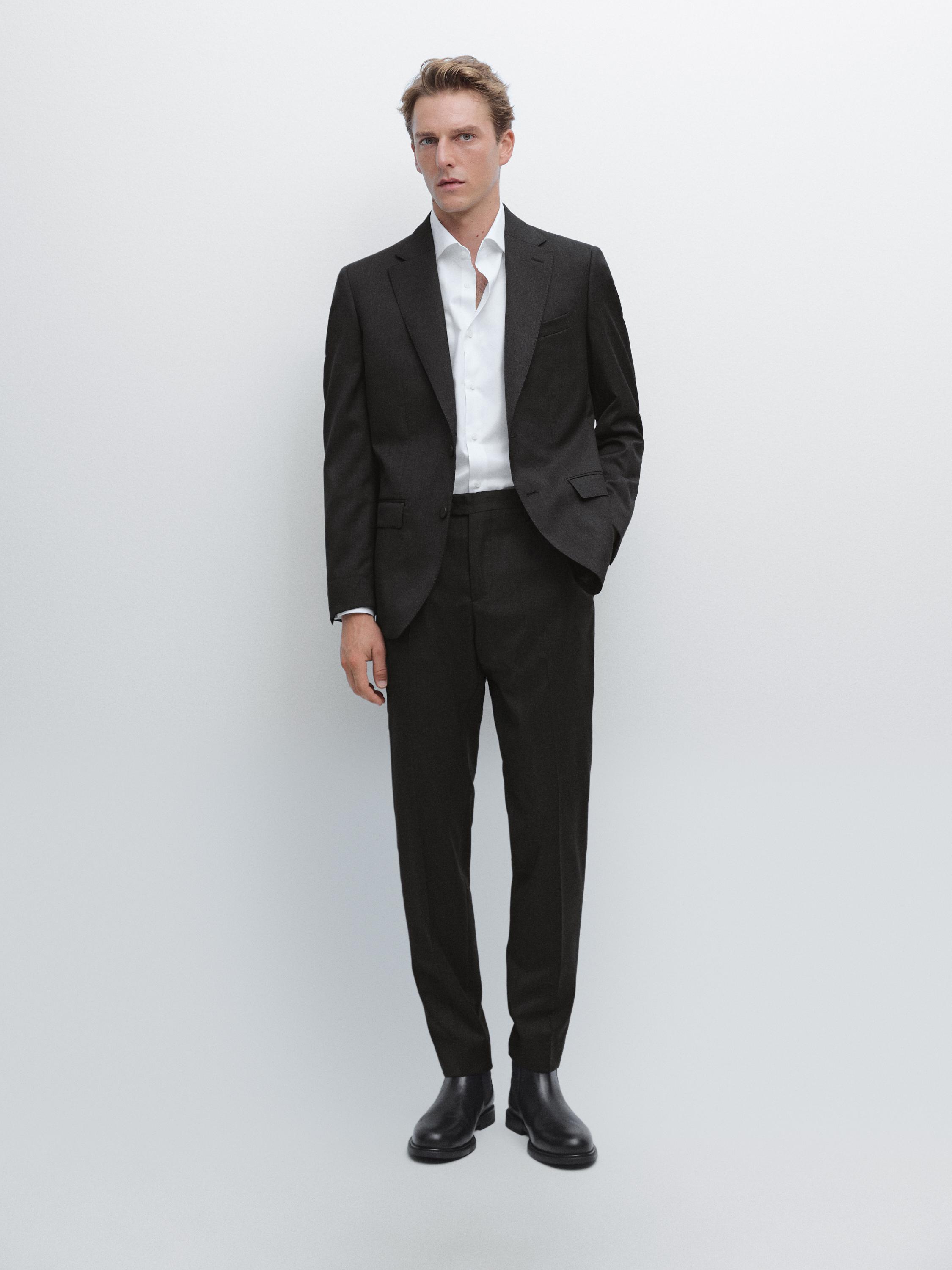 Conjunto de traje marengo con americana de dos botones, camisa blanca, pantalón recto y botas negras. Bolsillos laterales visibles.