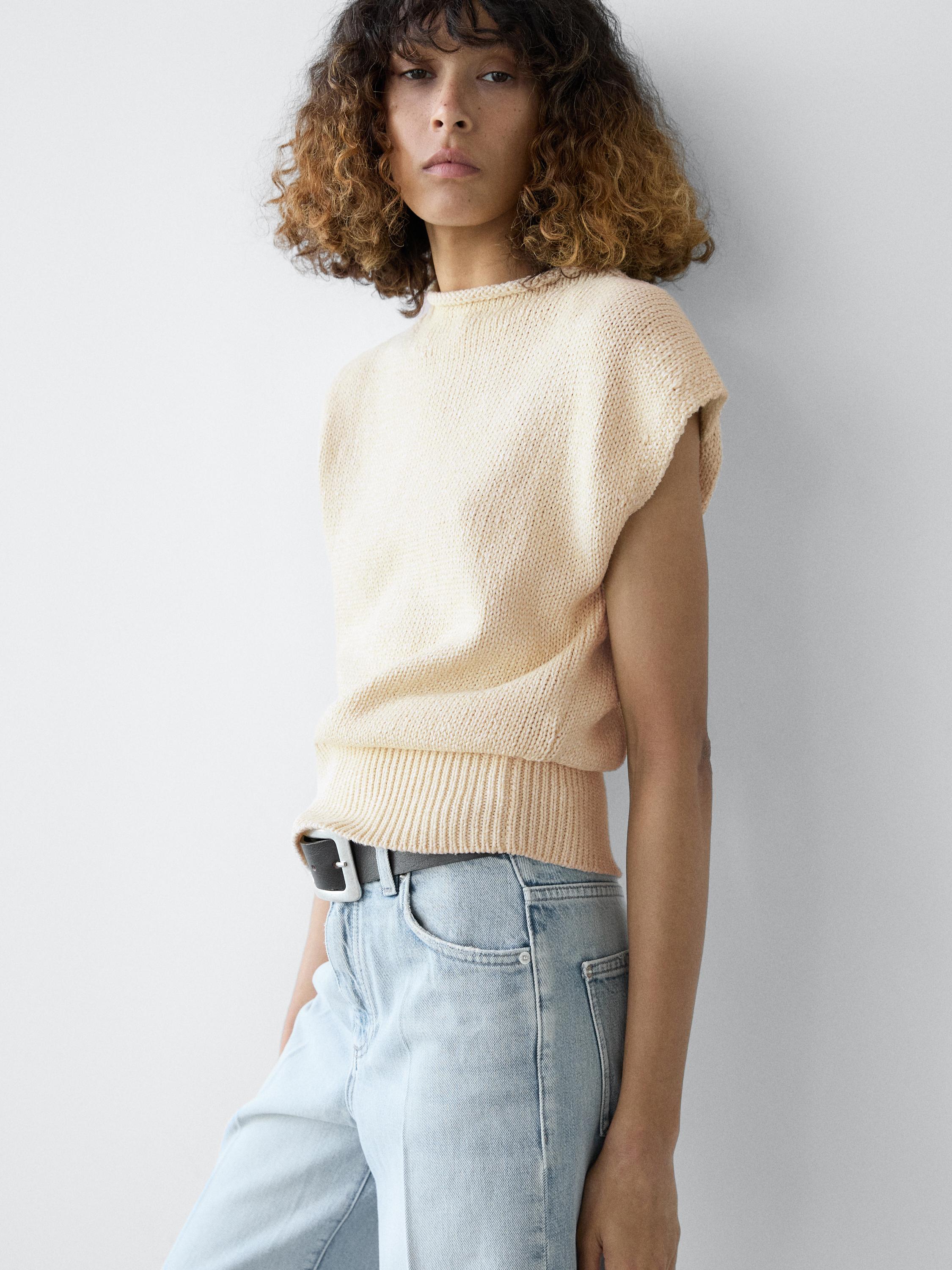 Plan moyen de gilet écru sans manches, col rond et coupe ample, associé à un jean bleu clair et une ceinture noire.