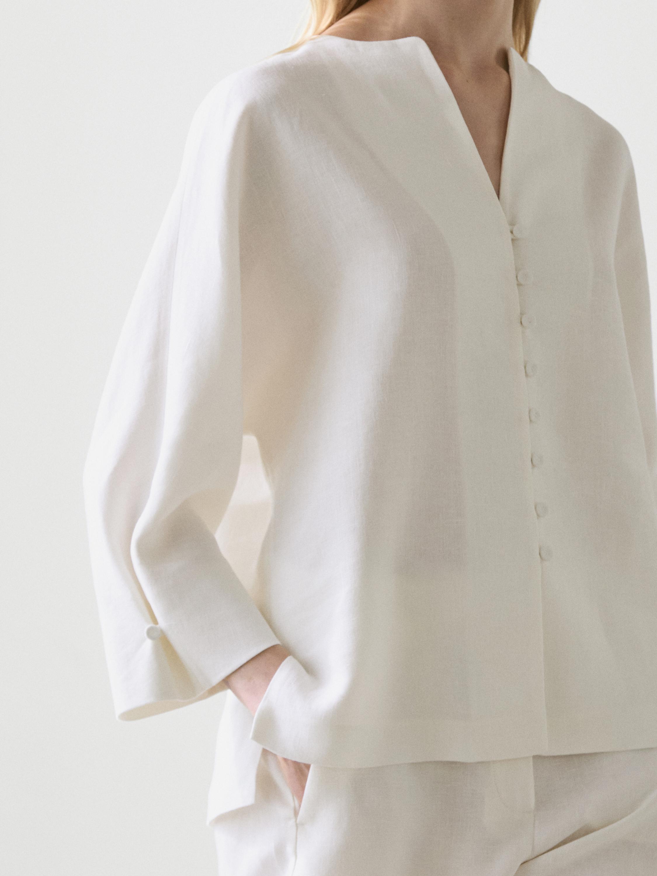 Plano medio de blusa blanca con botonadura frontal y mangas largas, detalle de bolsillo lateral.