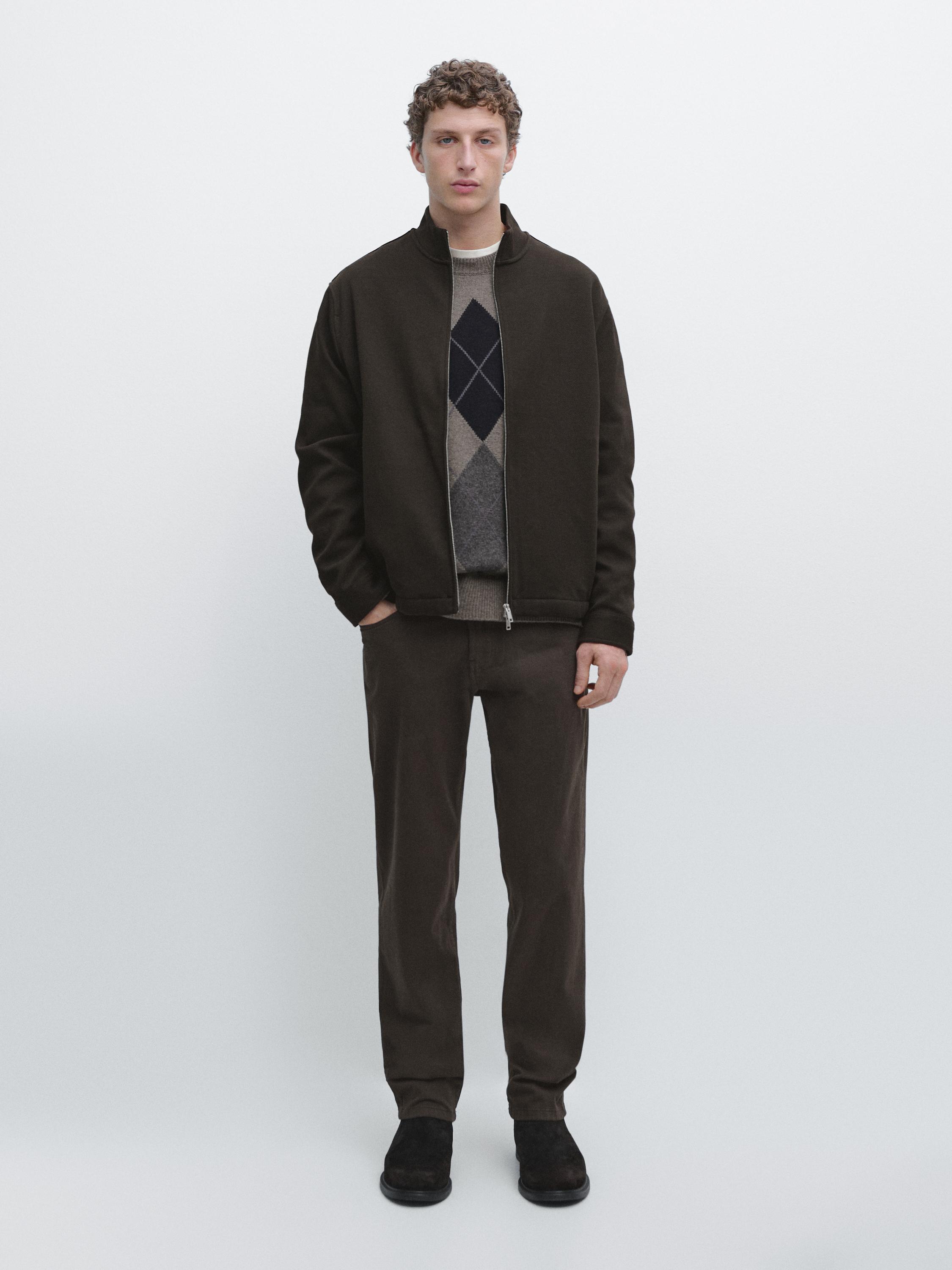 Conjunto de casaco castanho com fecho éclair, camisola com losangos e calças castanhas.