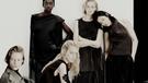 Cinq mannequins posent en vêtements sombres dans un environnement minimaliste. Une mannequin porte de longs gants noirs. L'image a un ton doux.