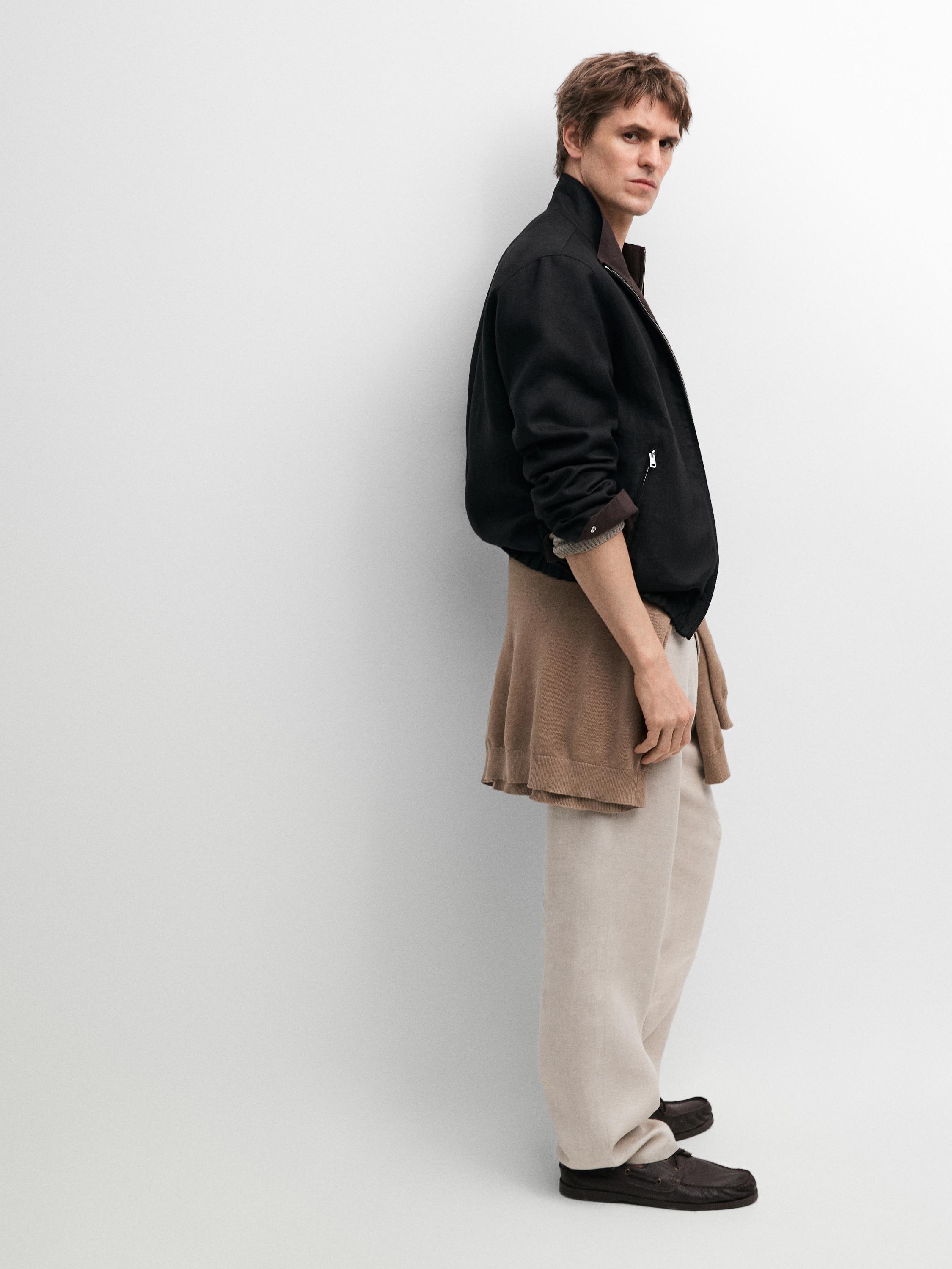 Conjunto de cazadora negra con cremallera lateral, pantalones beige amplios y suéter marrón atado a la cintura.