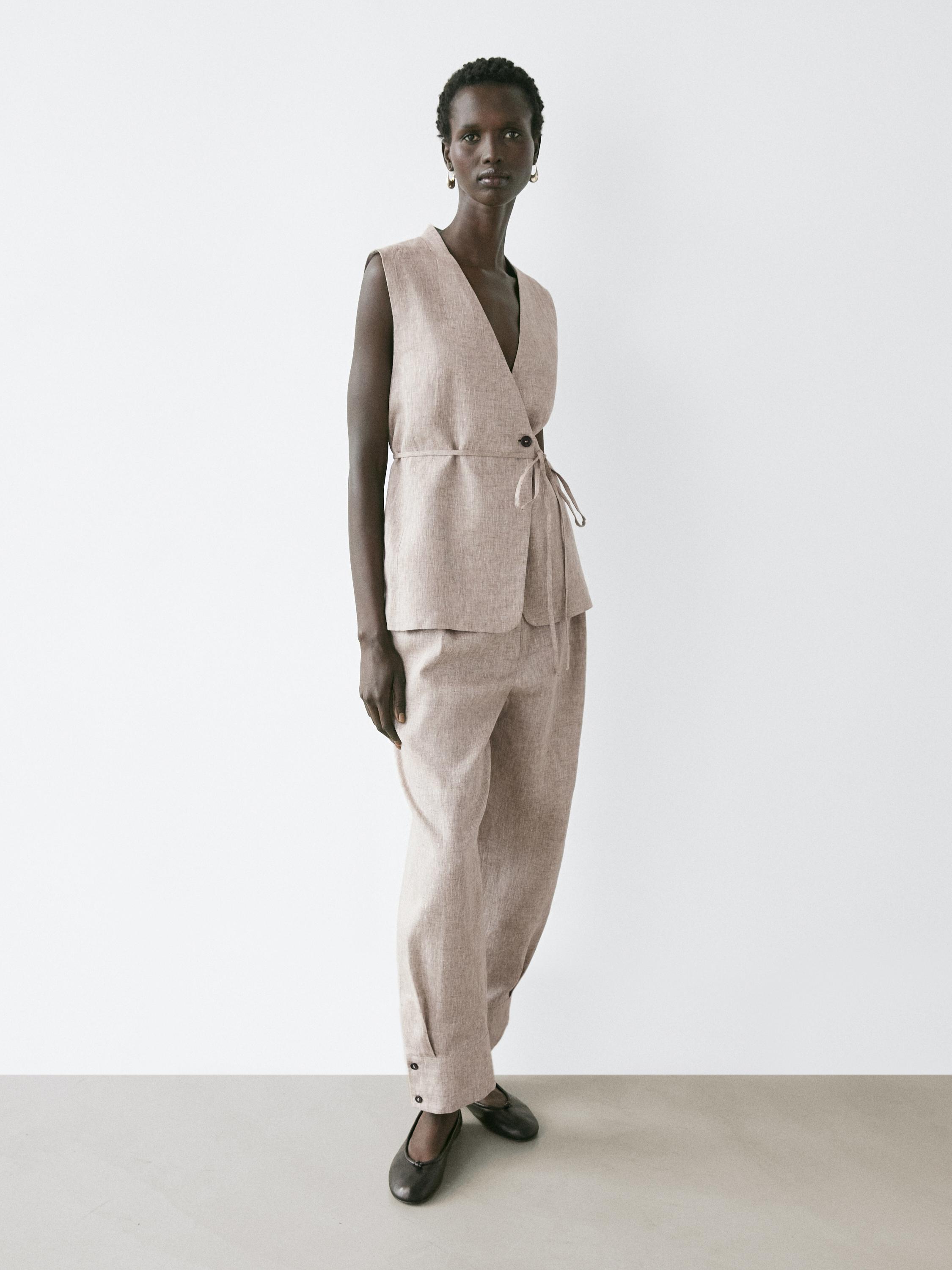 Бежевый комплект из жилета без рукавов с поясом и брюк в тон с пуговицами на щиколотках.