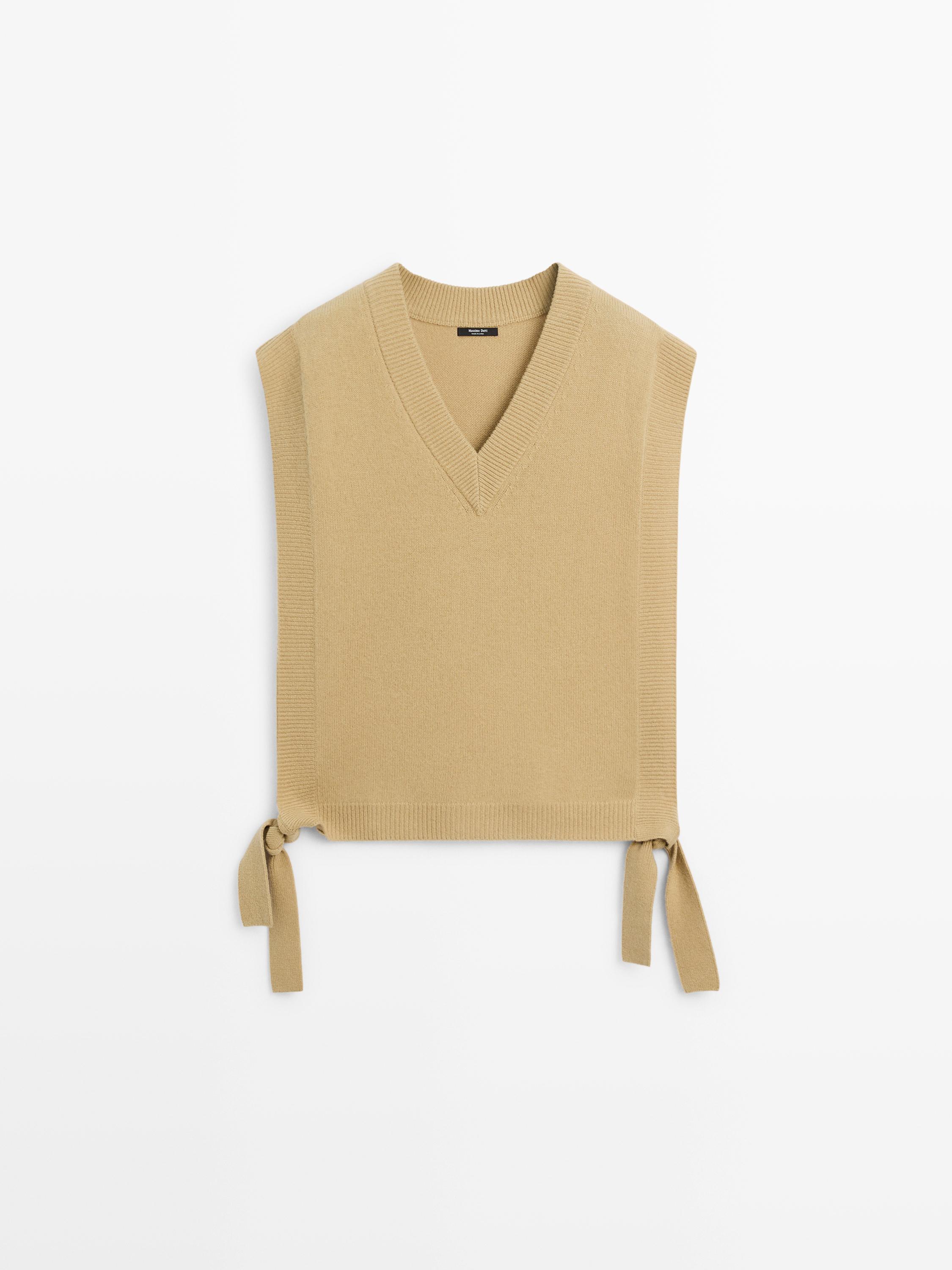 Vooraanzicht van een mouwloos beige gilet met V-hals en strikjes aan de zijkant.