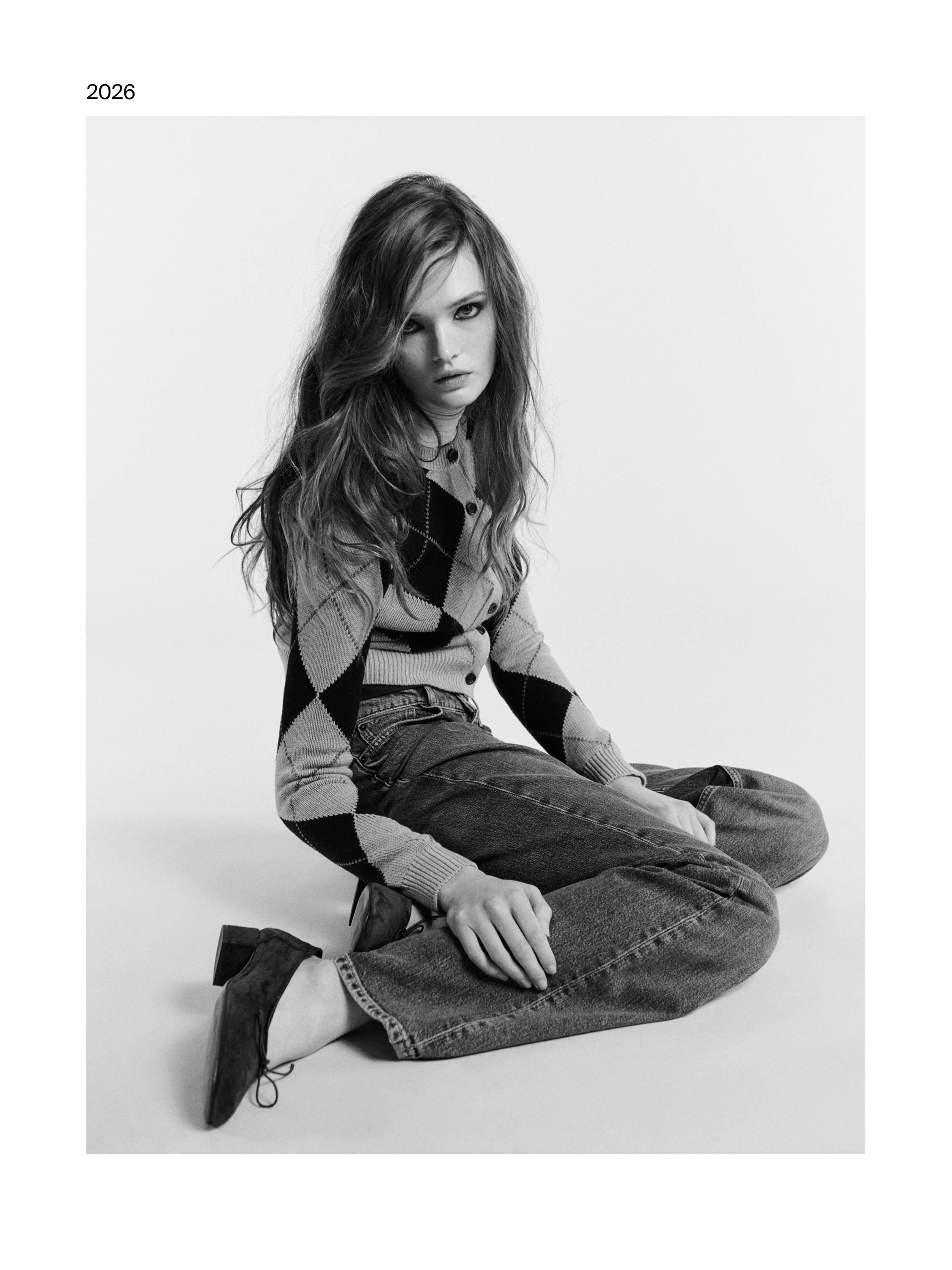 Rombo formos megztinis ir tiesūs džinsai. Tekstas "2026" viršutiniame kairiajame kampe. Žemakulniai bateliai.