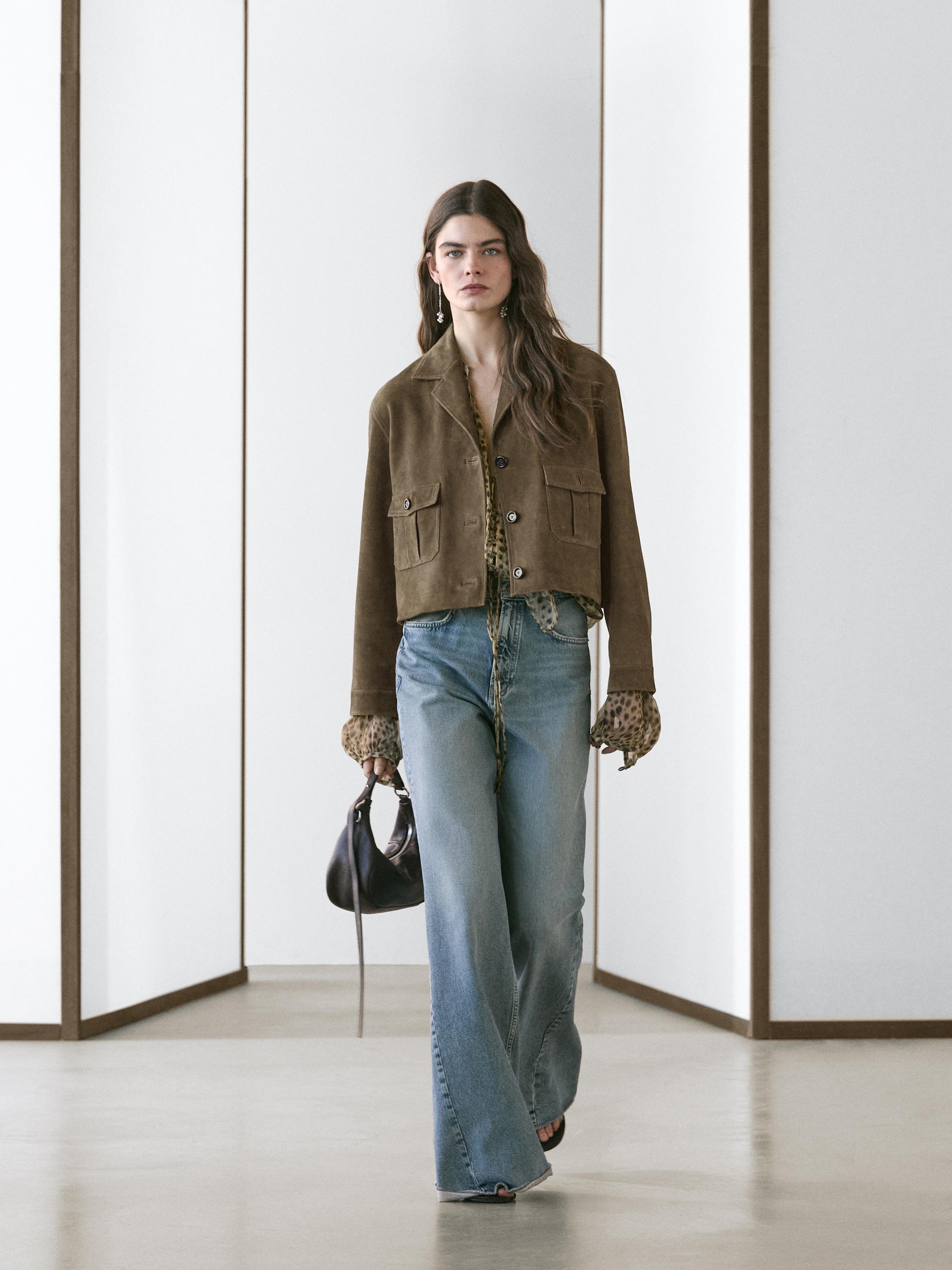 Conjunto de casaco curto cáqui com bolsos frontais e botões, jeans azuis de perna larga e luvas estampadas.