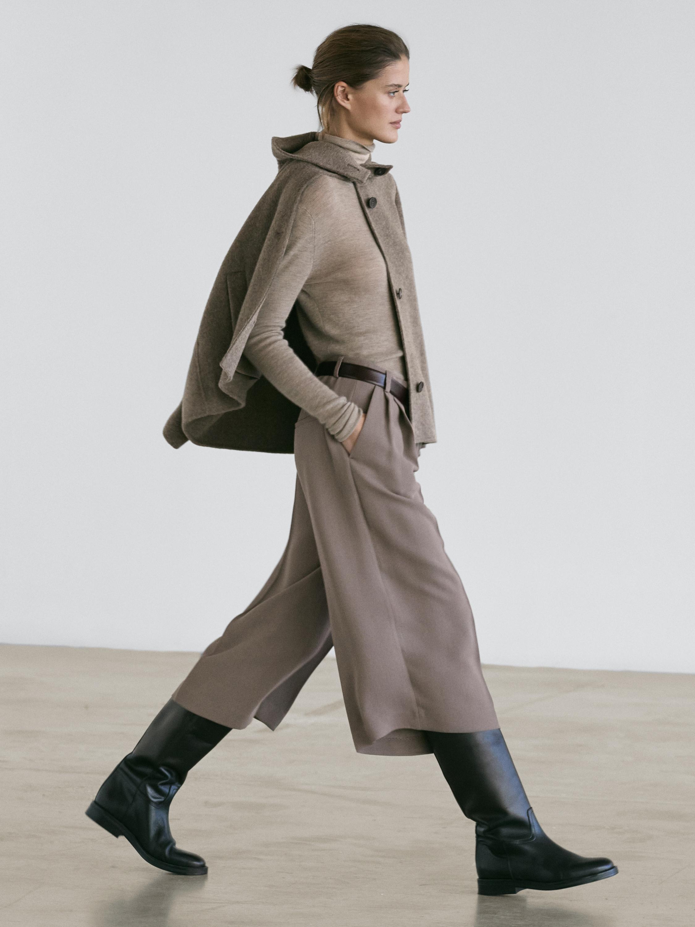 Komplet vključuje rjavkasto sivo pelerino z gumbi, pulover z visokim ovratnikom in dolge široke tekoče rjavkasto sive bermuda hlače, črni škornji.