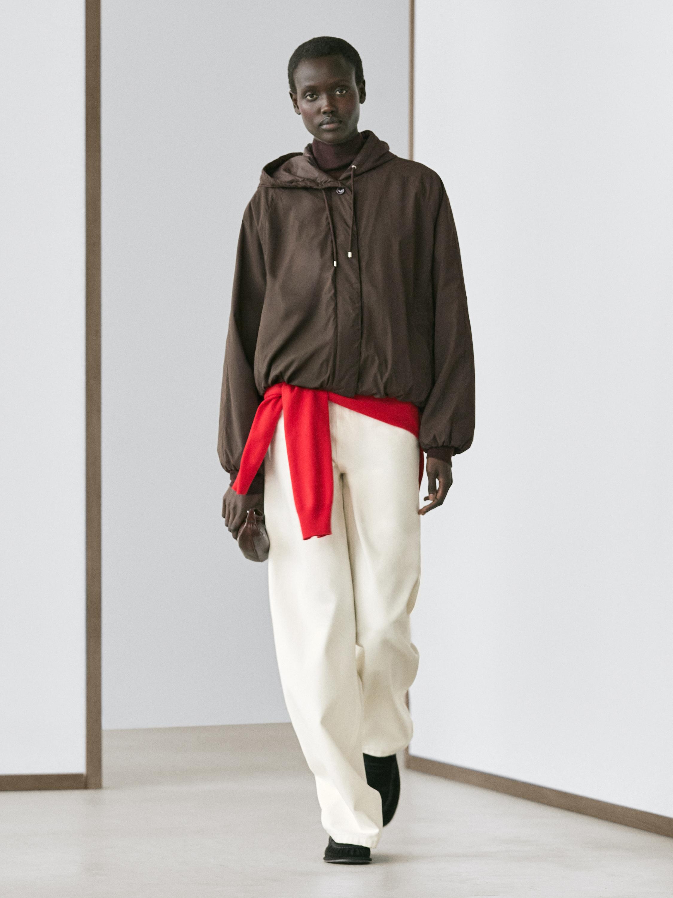 Zestaw składający się z czekoladowej puchowej kurtki z wysokim kołnierzem, białych szerokich spodni i czerwonego szalika zawiązanego w pasie.