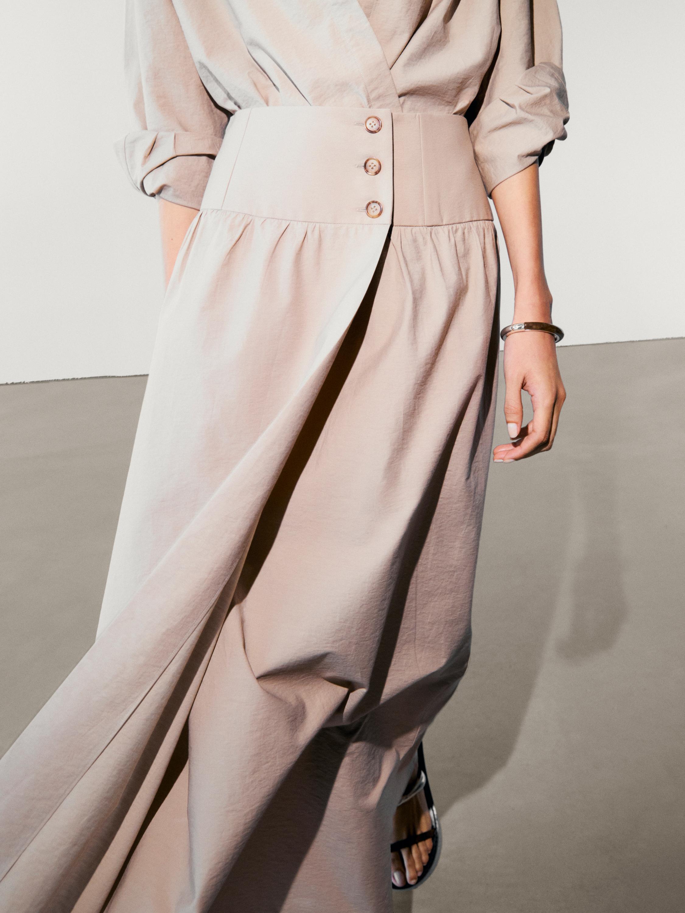 Détail de la jupe beige avec une large ceinture et des boutons, chemisier assorti avec des manches retroussées.