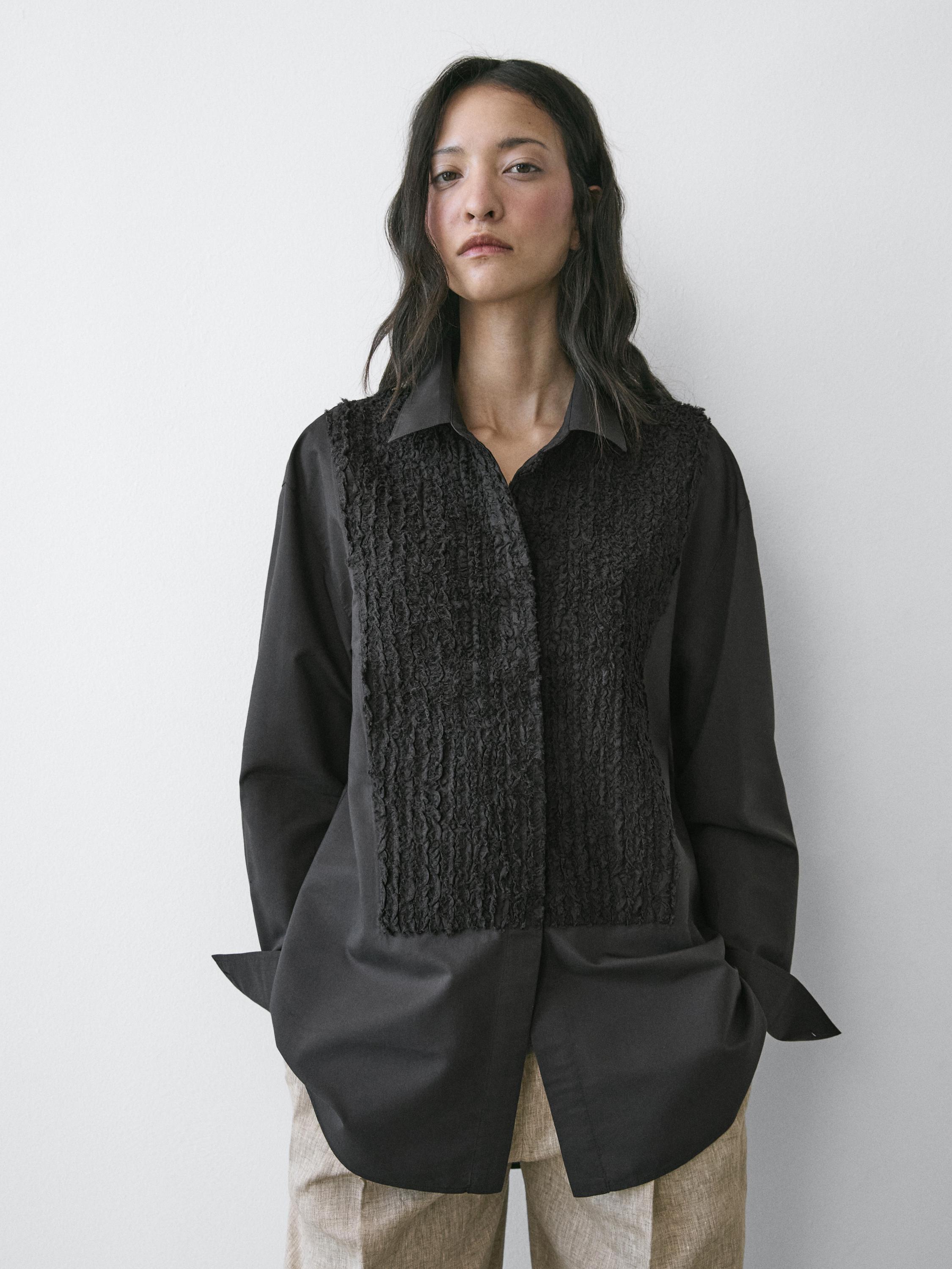 Komplet këmishë e zezë me jakë të qëndisur dhe xhepa anësorë, e kombinuar me pantallona bezhë.