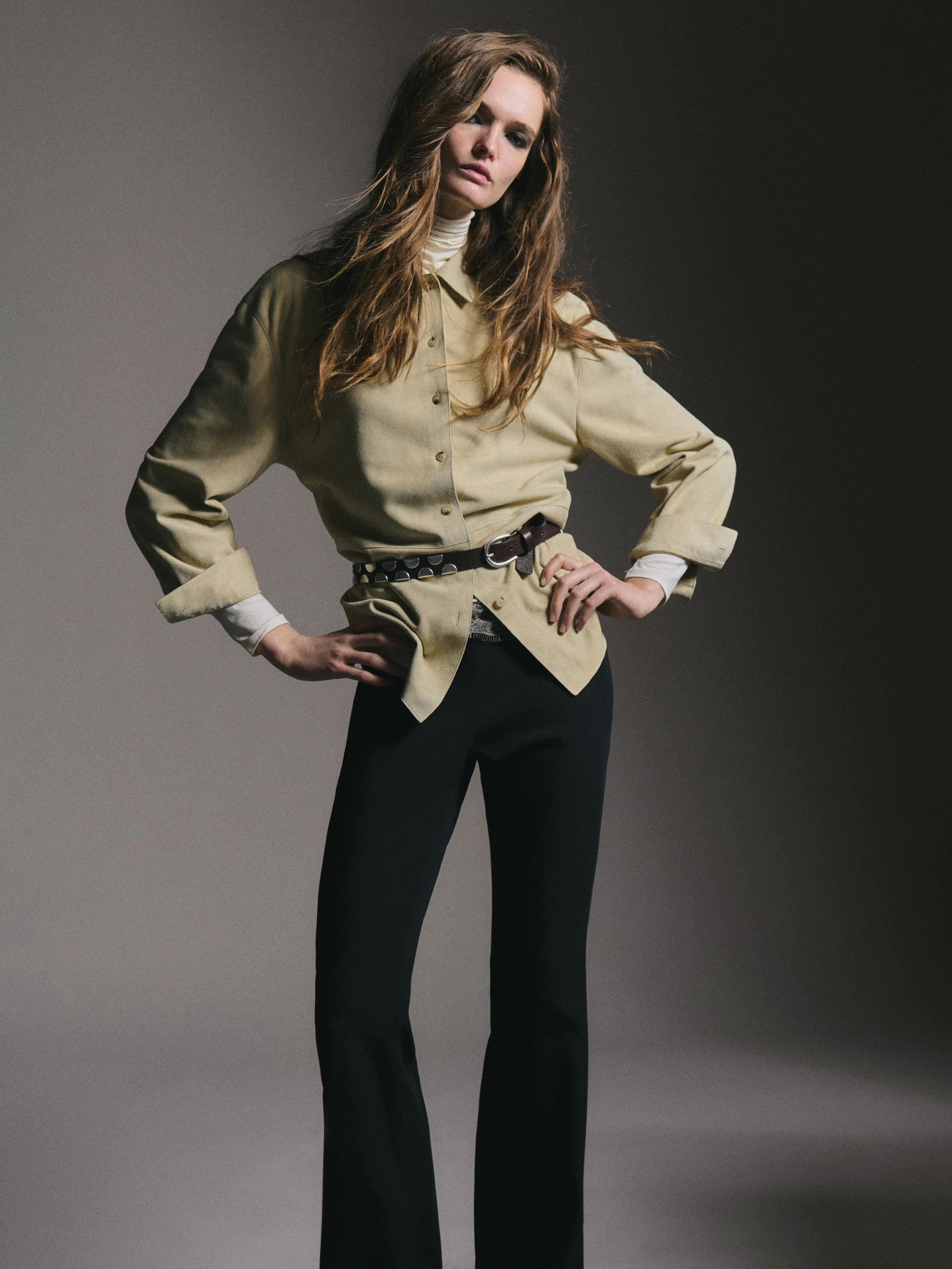 Smėlio spalvos marškinių su sagomis ir juodų platėjančių kelnių komplektas. Matomas tamsus diržas.