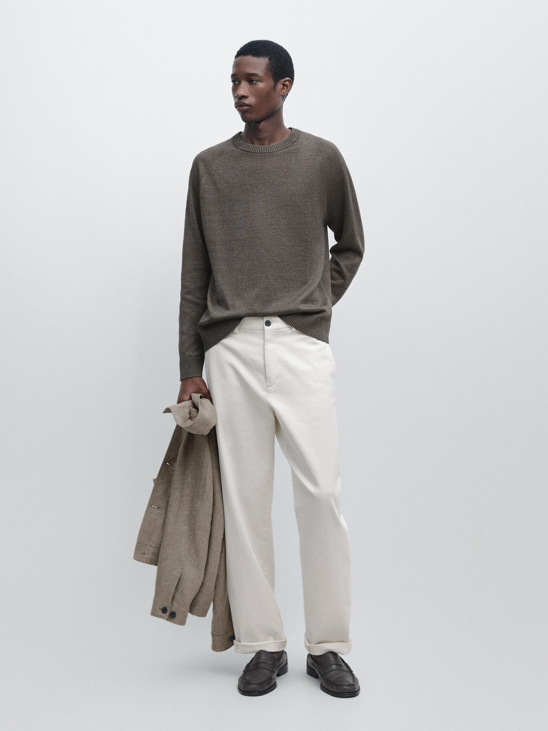 Set aus graugrünem Pullover und weißer Hose mit Falten. Er/Sie trägt dunkle Schuhe und hält einen beigen Mantel.