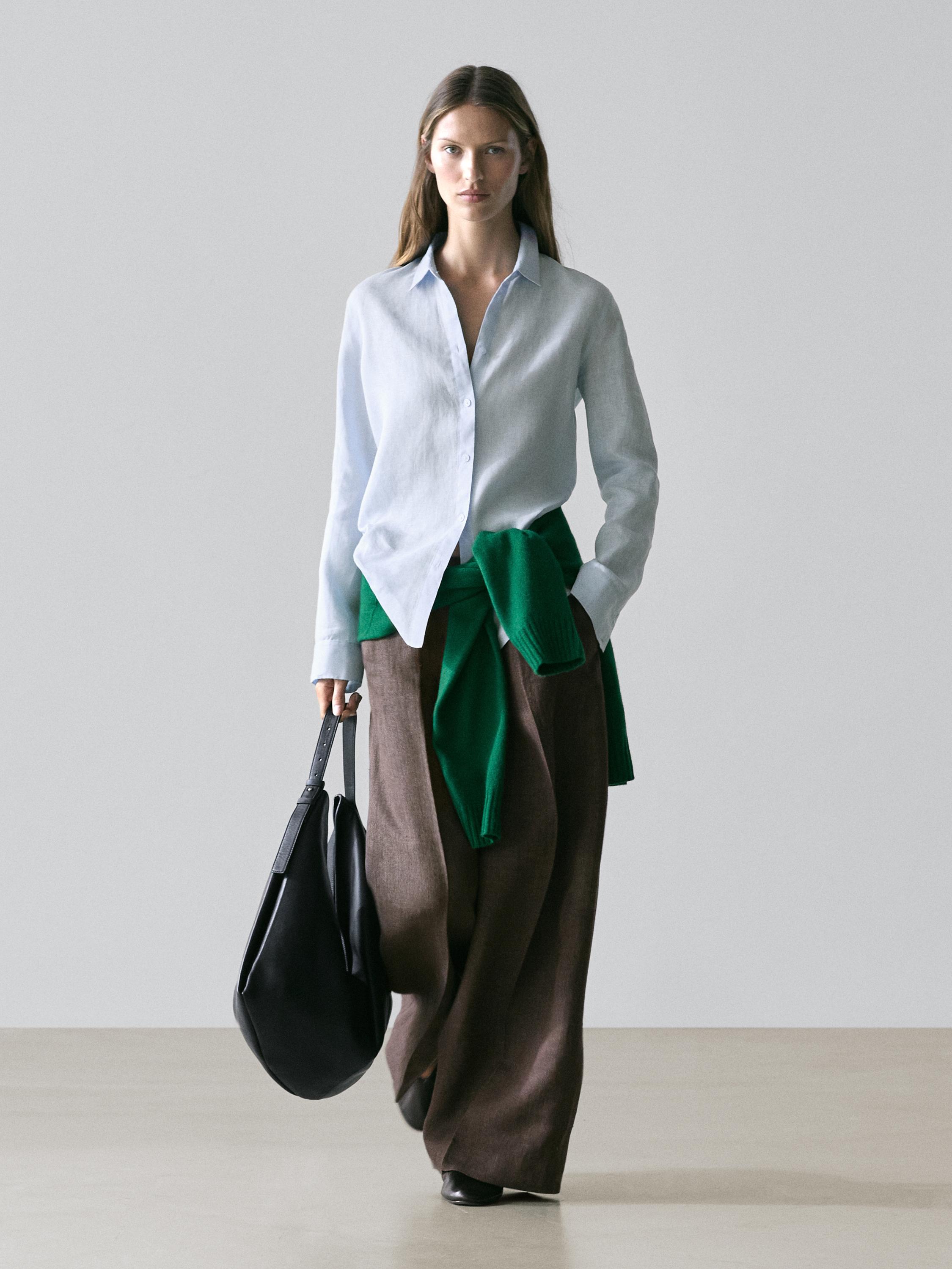 Conjunto de camisa celeste, pantalones marrones anchos y suéter verde atado a la cintura, bolso negro en mano.