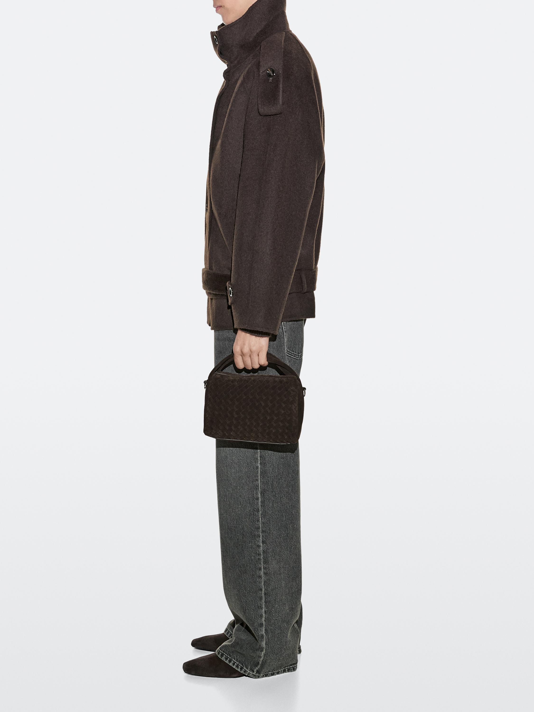 Conjunto de chaqueta marrón con cuello alto, pantalones grises y bolso pequeño marrón trenzado con asa superior.