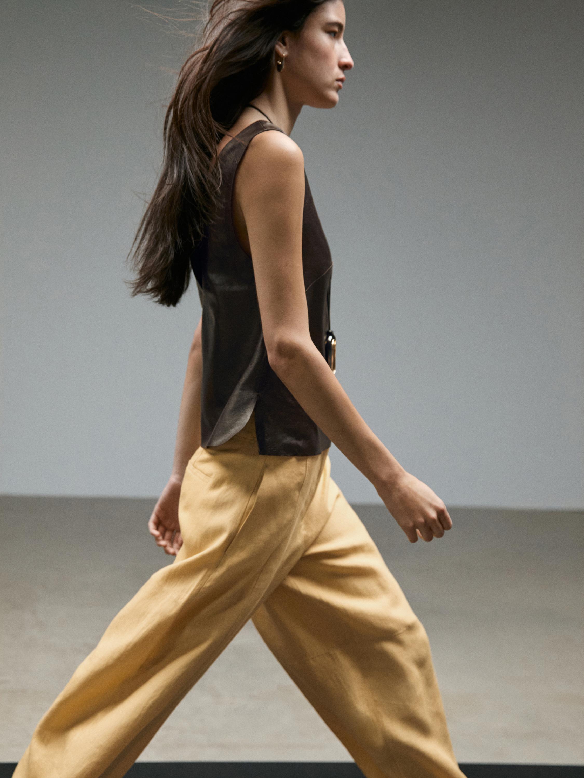 Plano medio de pantalón amarelo de perna ancha con pregos frontais e top sen mangas marrón escuro.