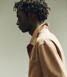 رجل بملف جانبي بشعر مجعد، يرتدي سترة بنية فاتحة. خلفية محايدة، أسلوب بسيط ومسترخٍ.