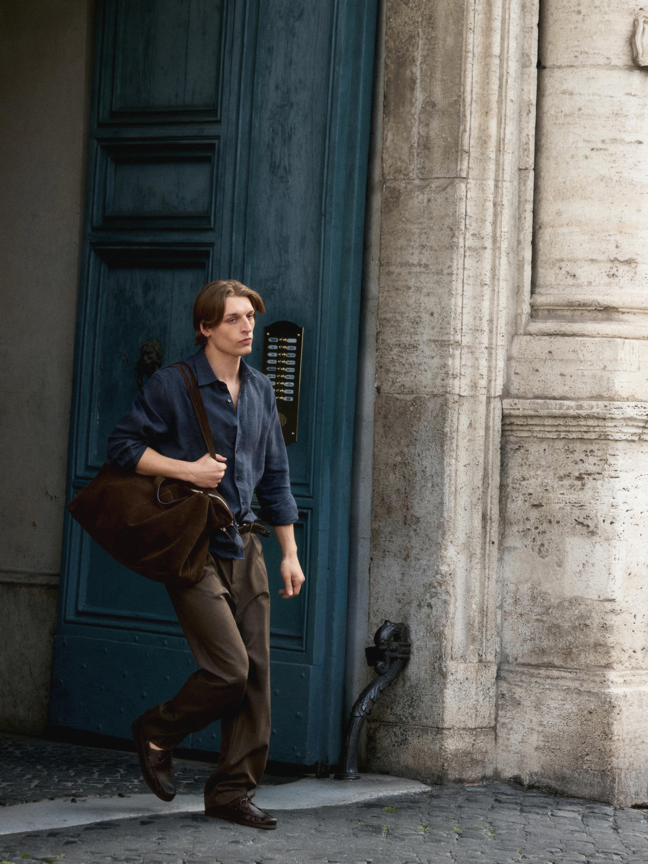 Koyu mavi gömlek ve kahverengi pantolon takımı. Omuzunda büyük, koyu kahverengi bir çanta taşıyor.