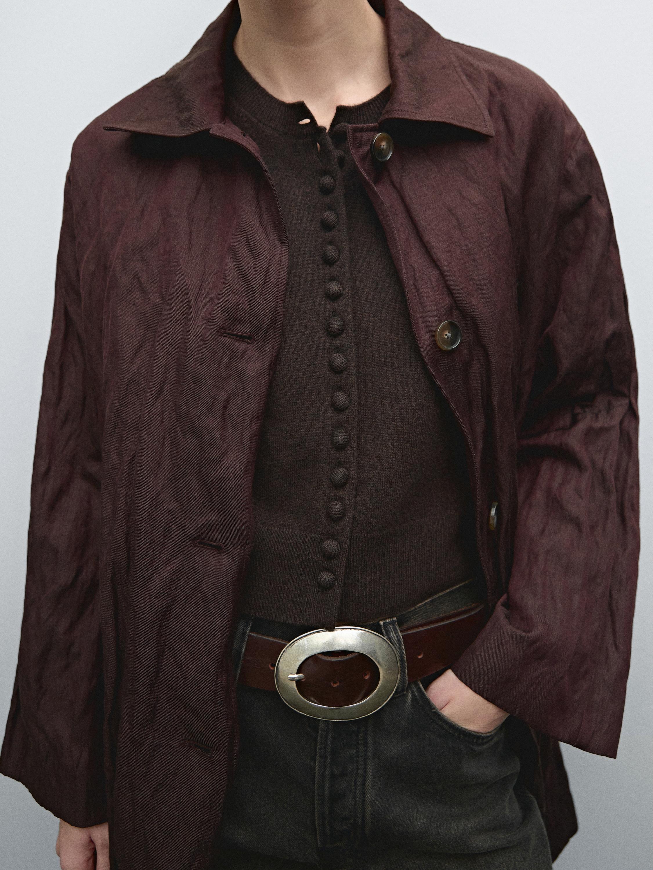 Conjunto de chaqueta marrón y suéter con botonadura, cinturón con hebilla circular y pantalones oscuros.