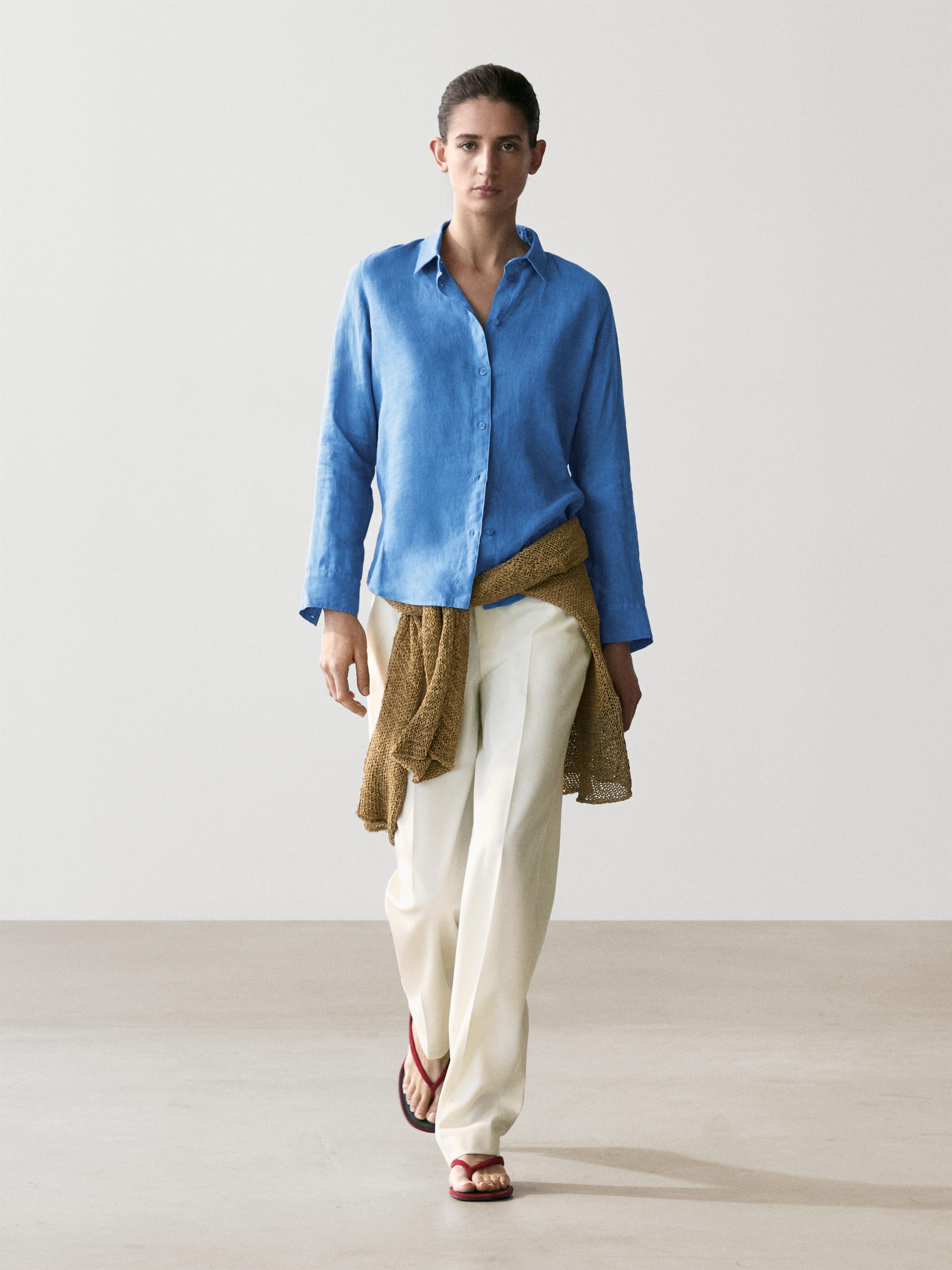 Ensemble composé d'une chemise bleu indigo boutonnée, d'un pantalon blanc ample et de sandales rouges. Pull marron noué à la taille.