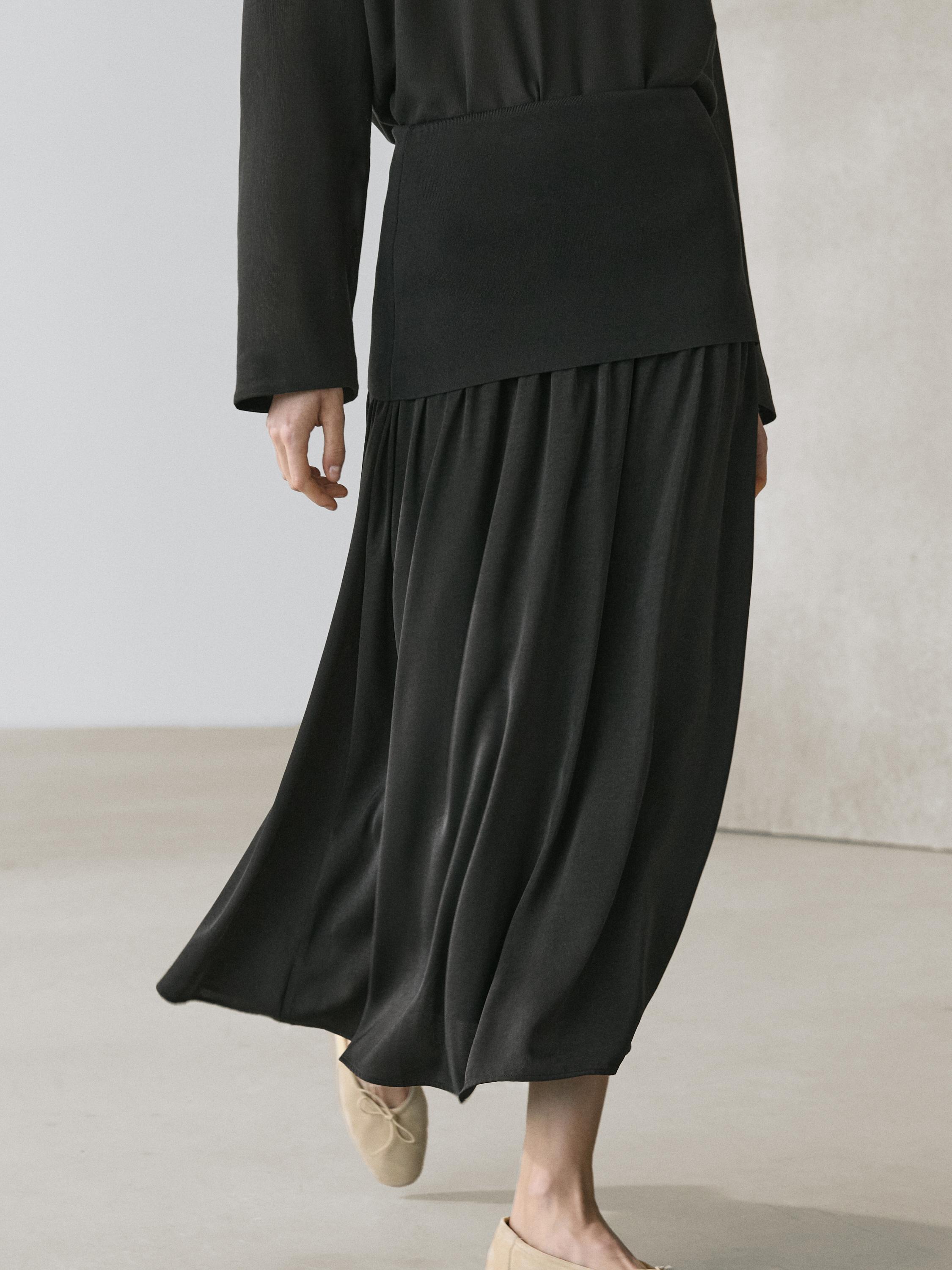 Middelgroot shot van een zwarte gebreide rok met brede taille en plooien, gecombineerd met een zwarte top met lange mouwen.