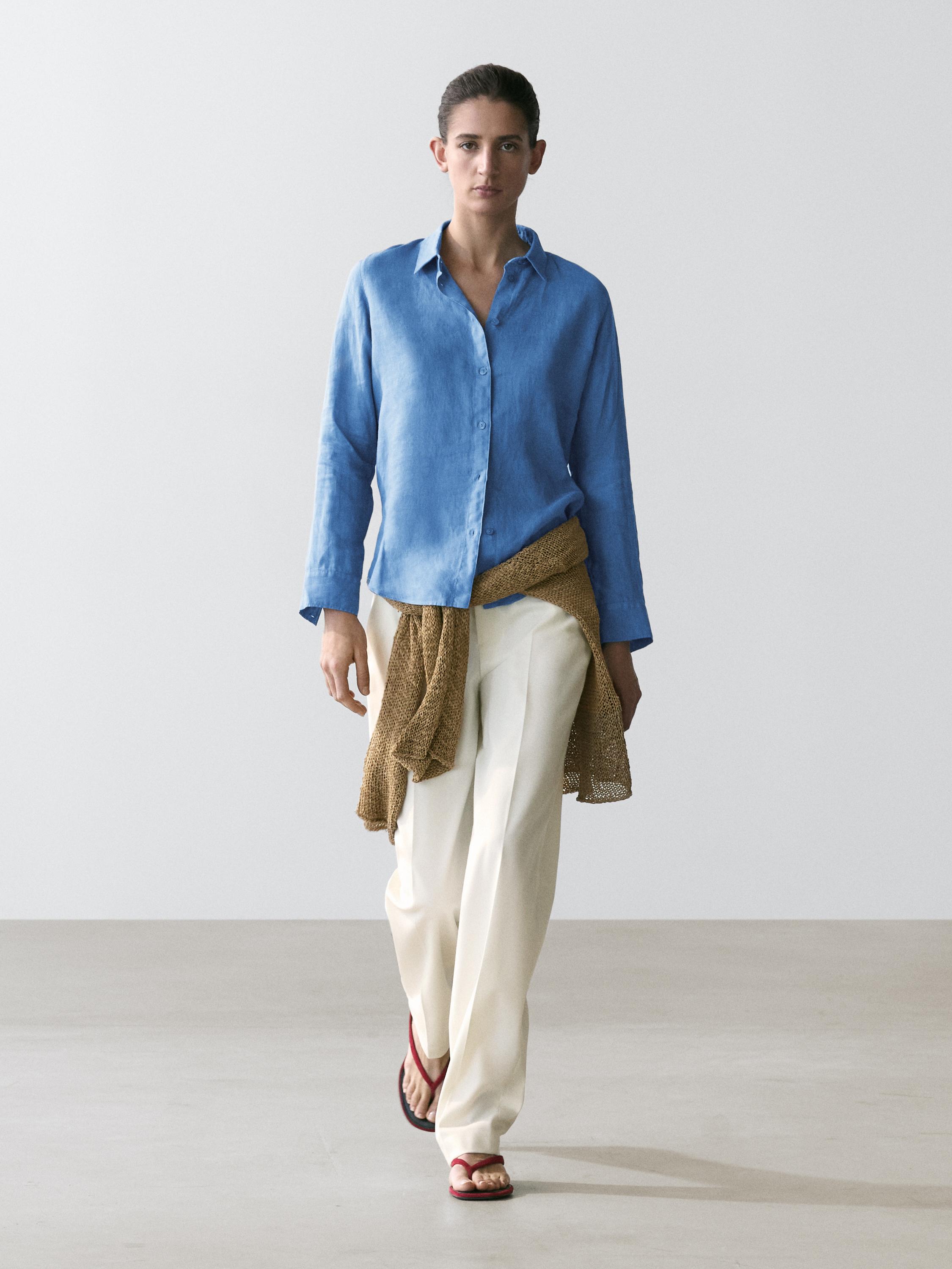 Fließendes indigoblaues Hemd, weite weiße Hose und rote Sandalen. Brauner Pullover um die Taille gebunden.