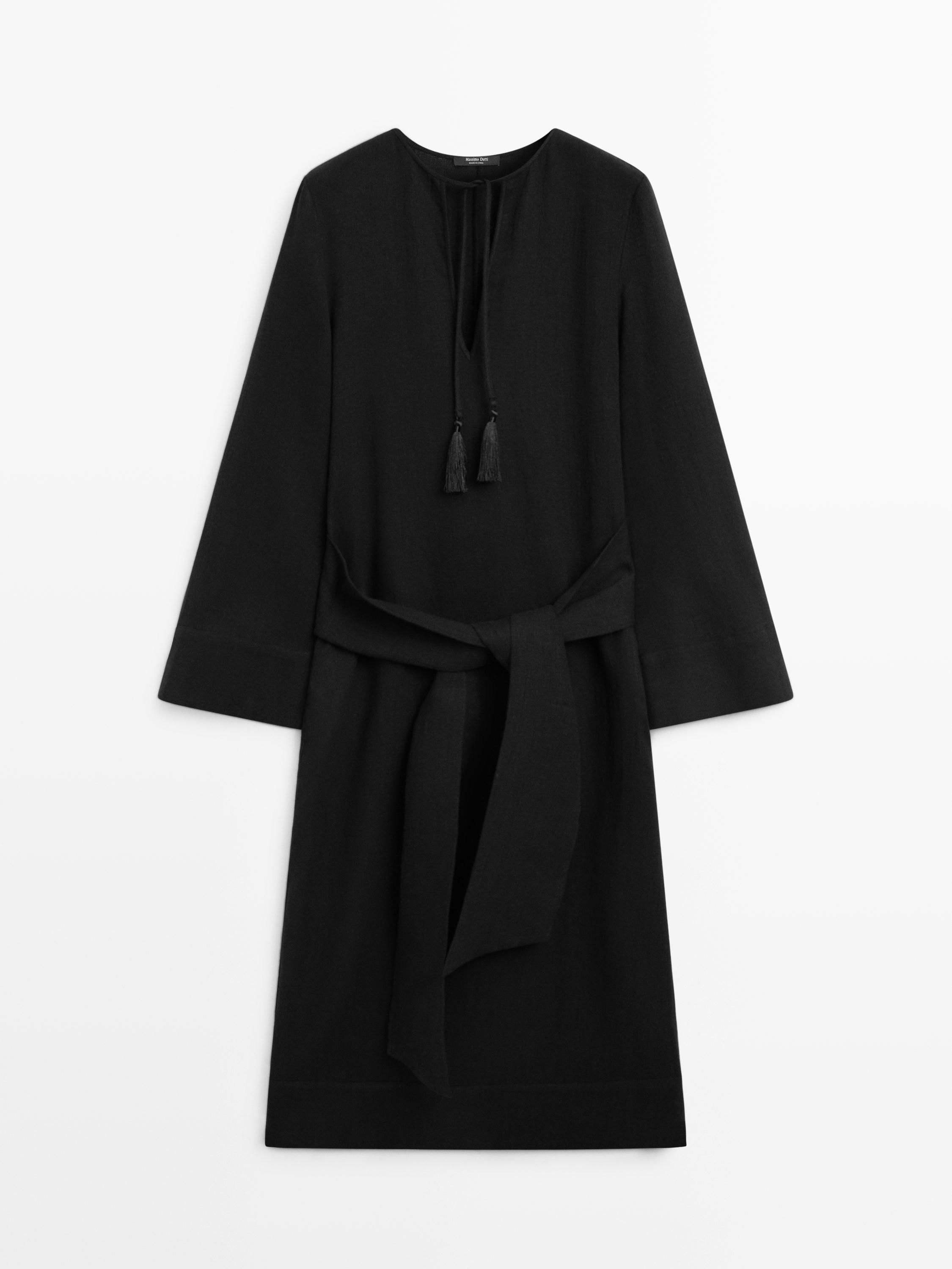 Vue de face de la robe noire avec un nœud à la taille, des manches longues et un décolleté avec des lacets et des glands.