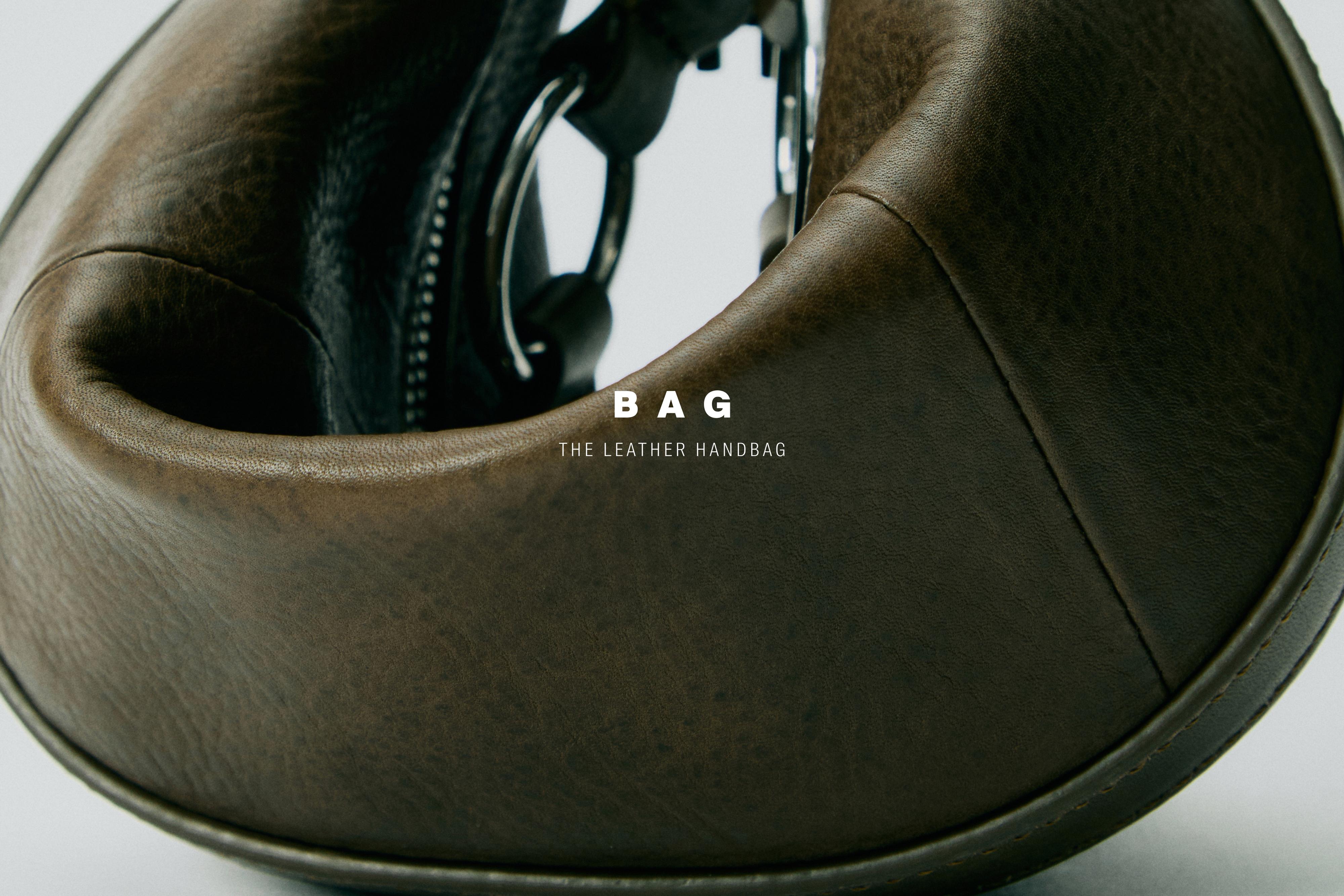 Detail einer khakifarbenen Tasche mit sichtbarem Text „BAG THE LEATHER HANDBAG“, Reißverschluss und gebogener Form.