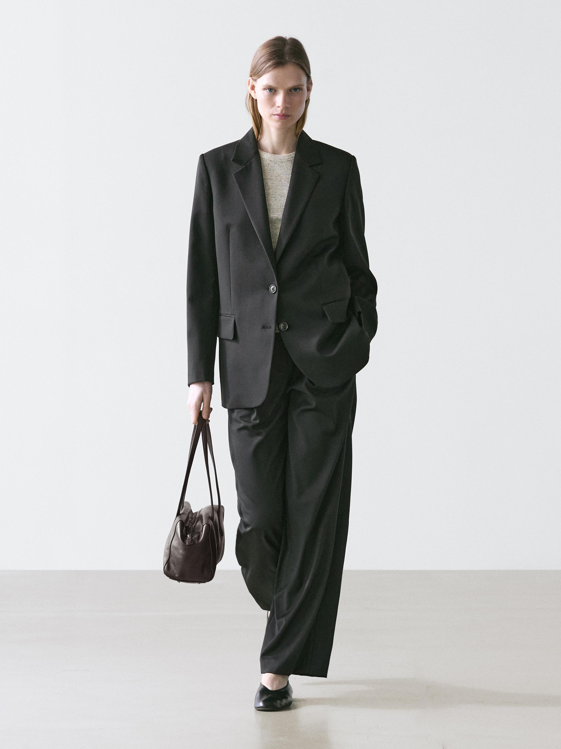 Ensemble de tailleur noir avec veste à deux boutons et pantalon large. Porte un sac marron et des chaussures noires.