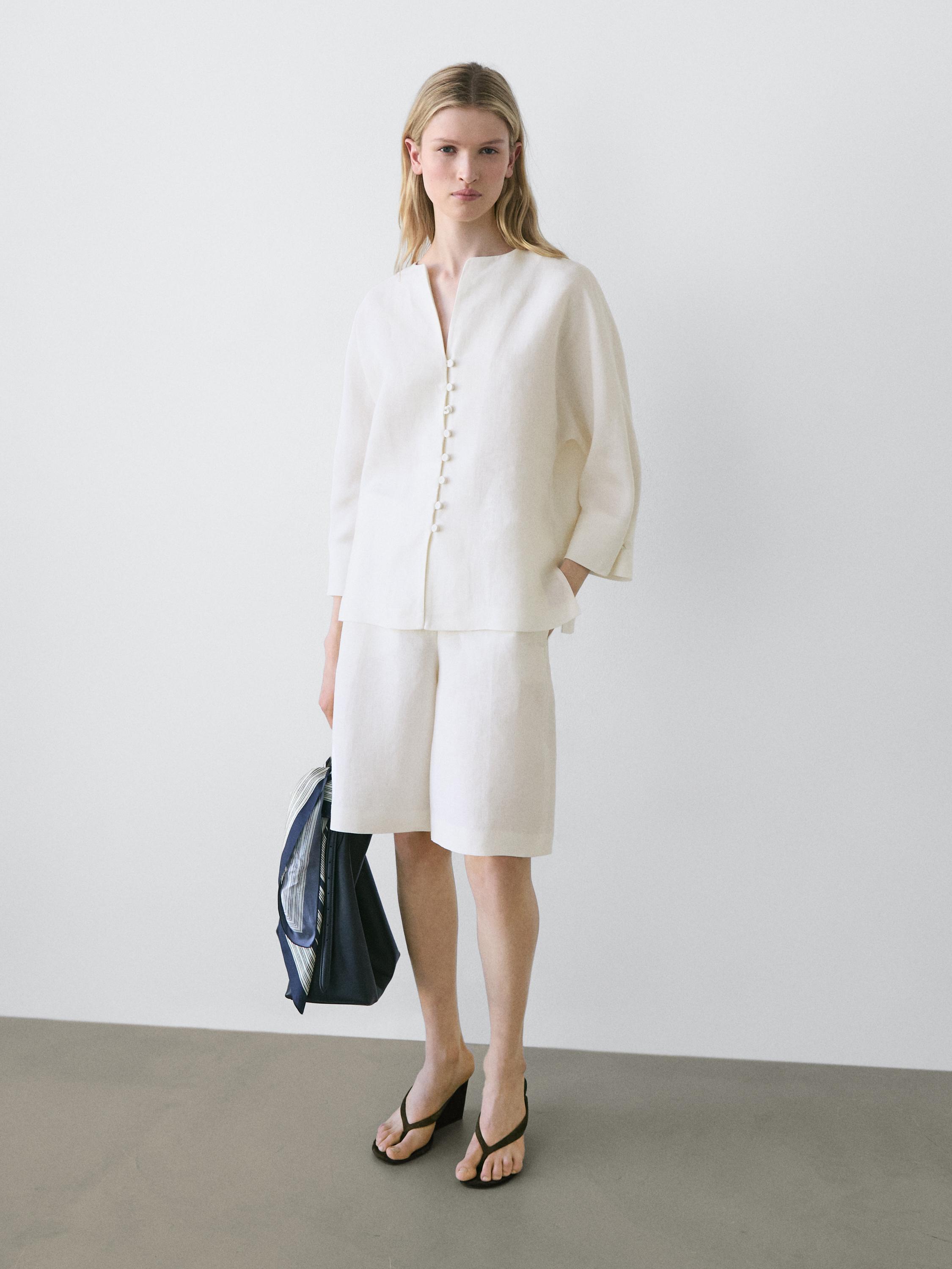 Conjunto de blusa blanca con botones frontales y bermudas a juego. Lleva sandalias negras y bolso azul.