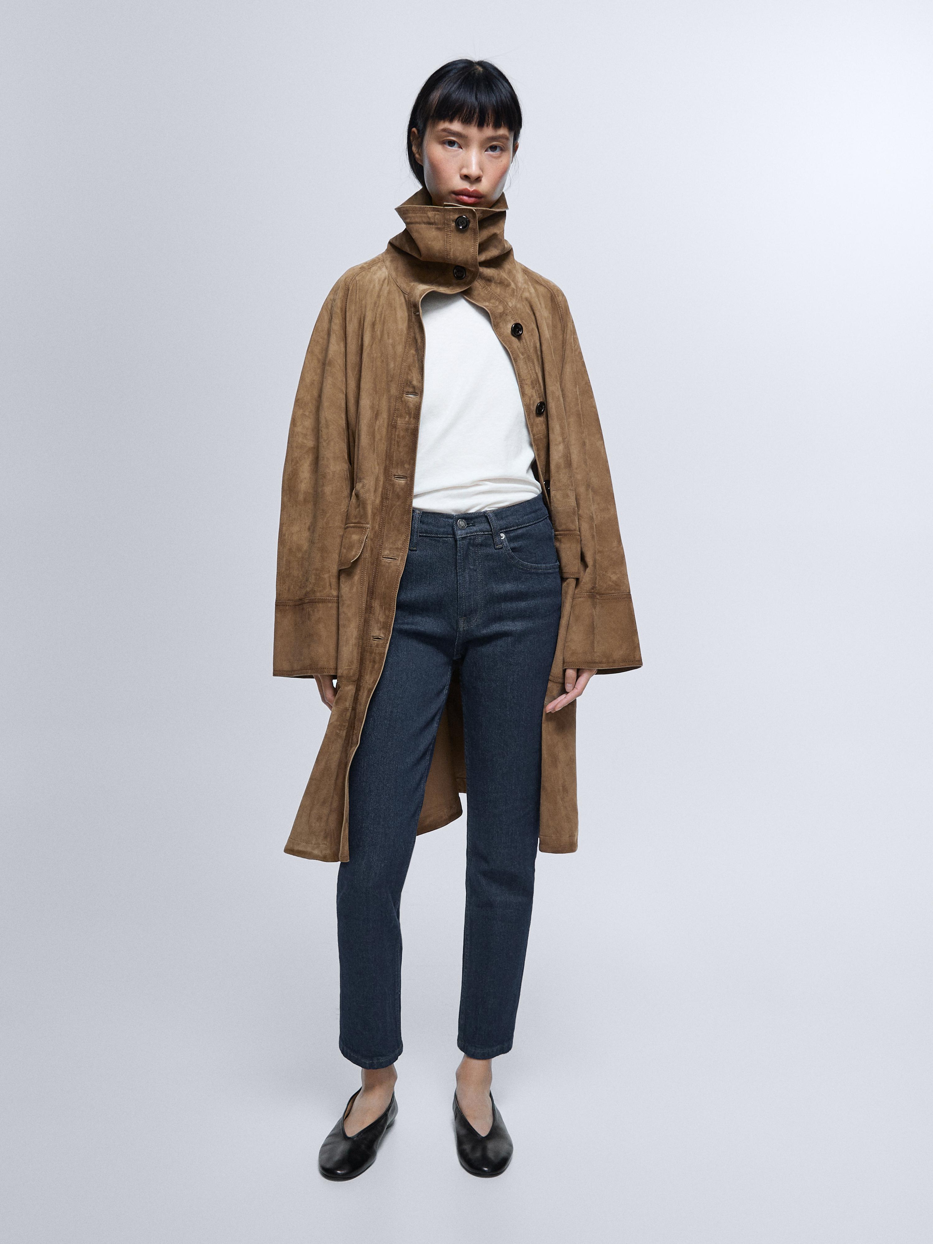 Conxunto de vaqueiros azul escuro slim fit tiro medio, abrigo marrón con cuello alto e botóns, camiseta branca e sapatos negros.