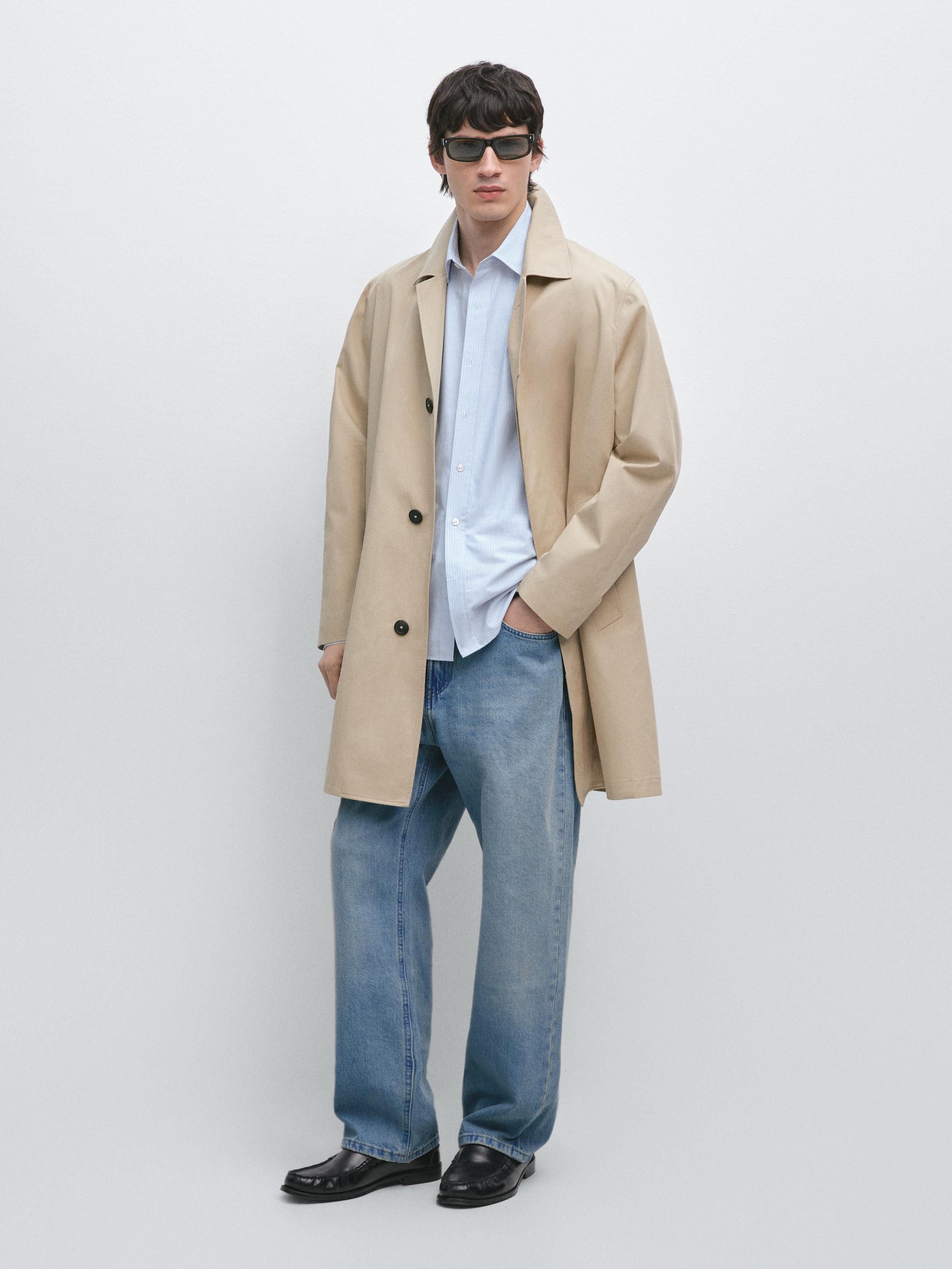 Siyah düğmeli bej trençkot takımı, açık mavi gömlek, geniş paça mavi kot pantolon ve siyah ayakkabılar.