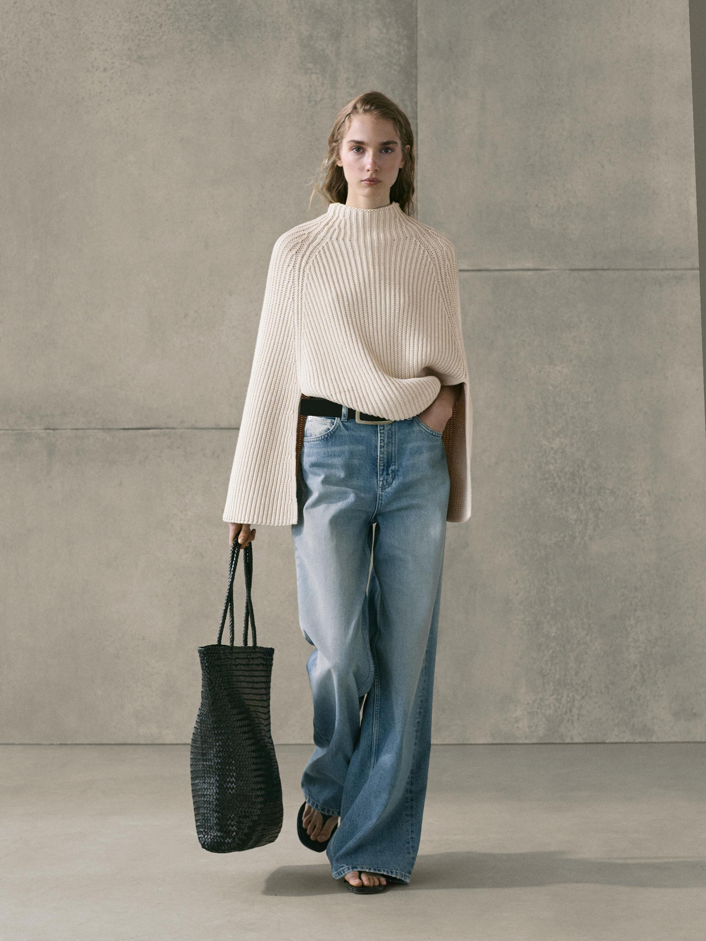 Conjunto de suéter beige de punto y jeans azul claro de pierna ancha con tiro alto, bolso negro tejido y sandalias.
