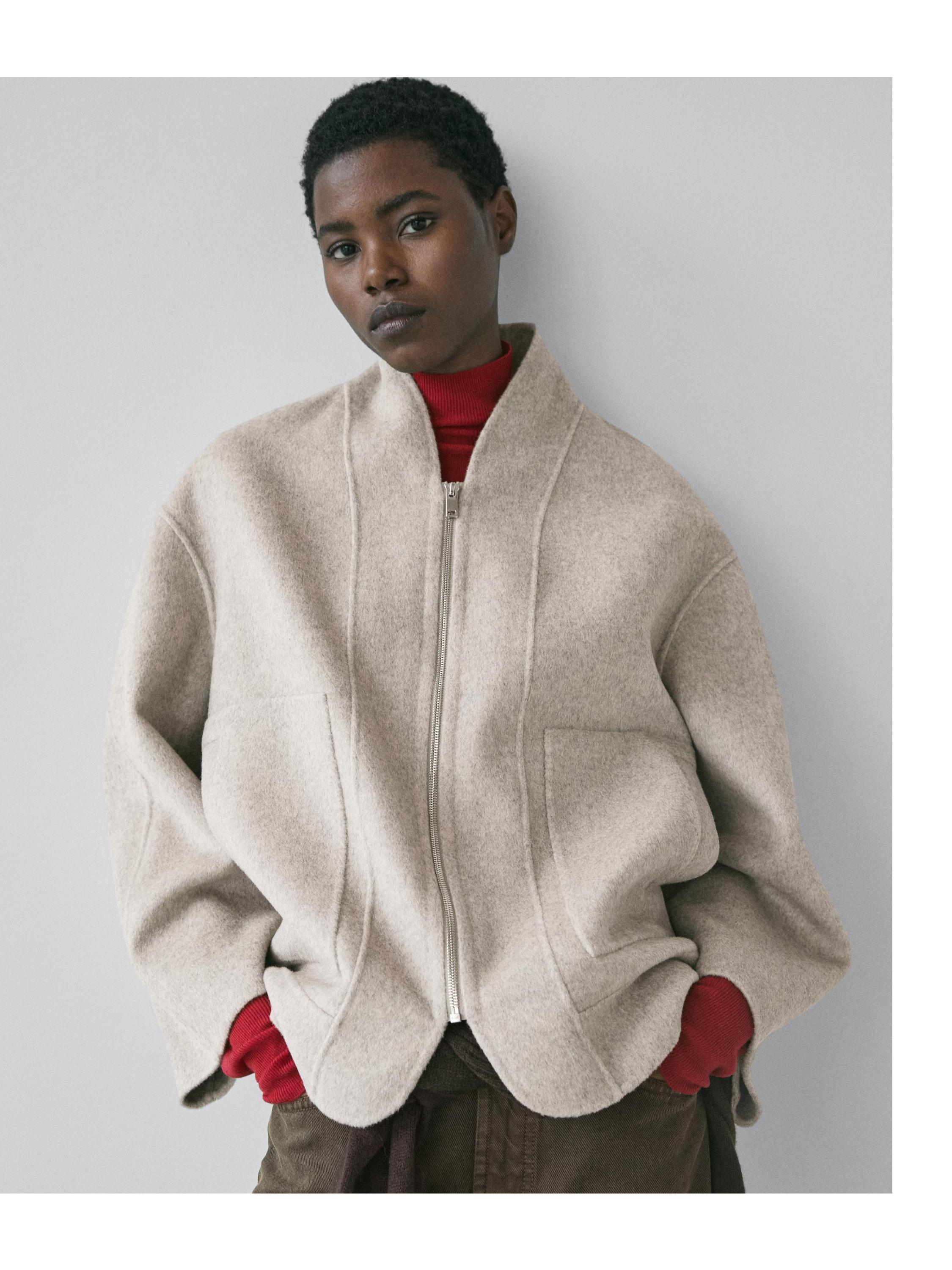 Plano medio de abrigo gris perla con cierre y bolsillos laterales, sobre suéter rojo visible en puños.