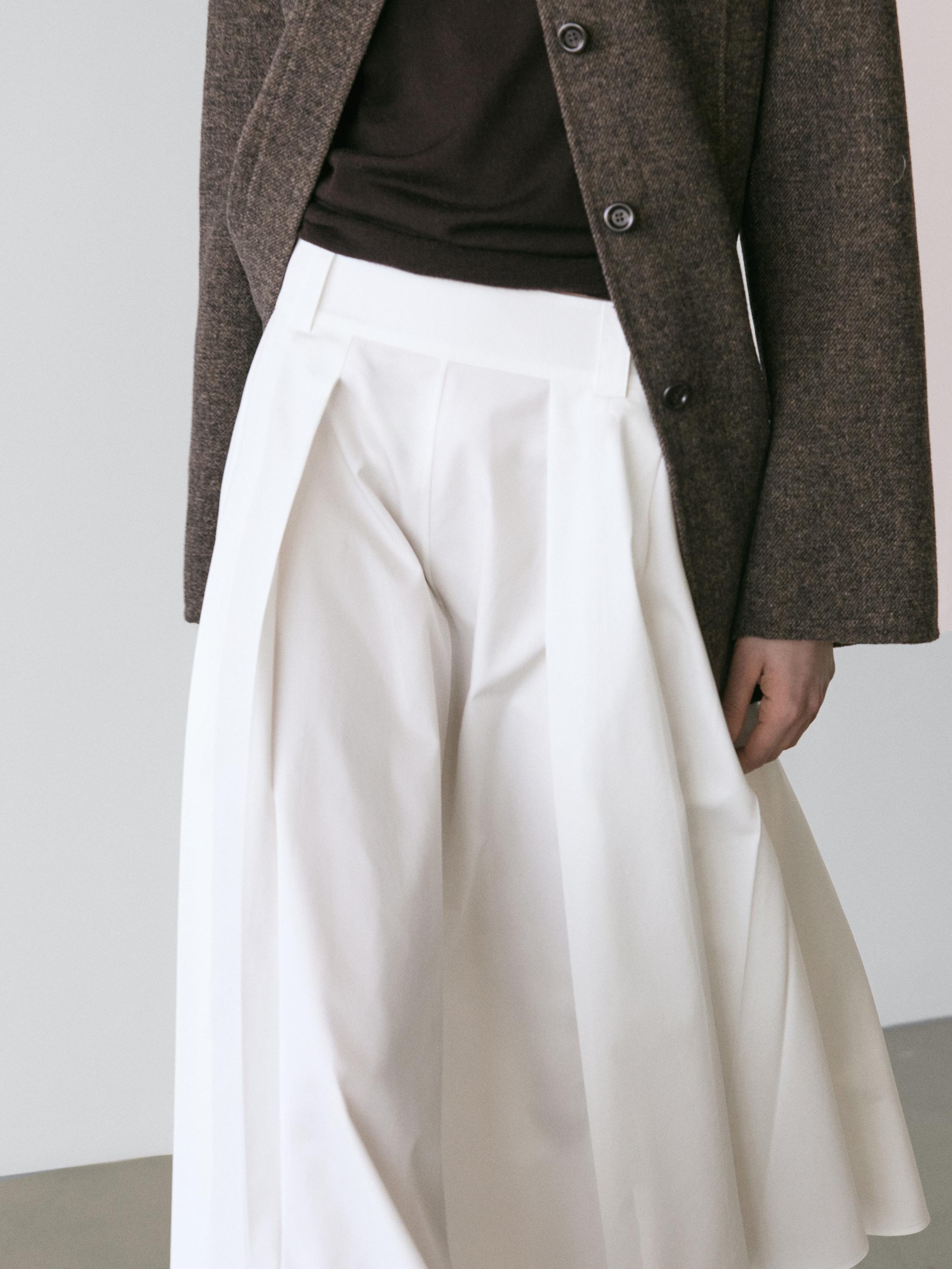 "Plano medio de falda midi blanca con detalle de trabillas y pliegues, combinada con chaqueta marrón."