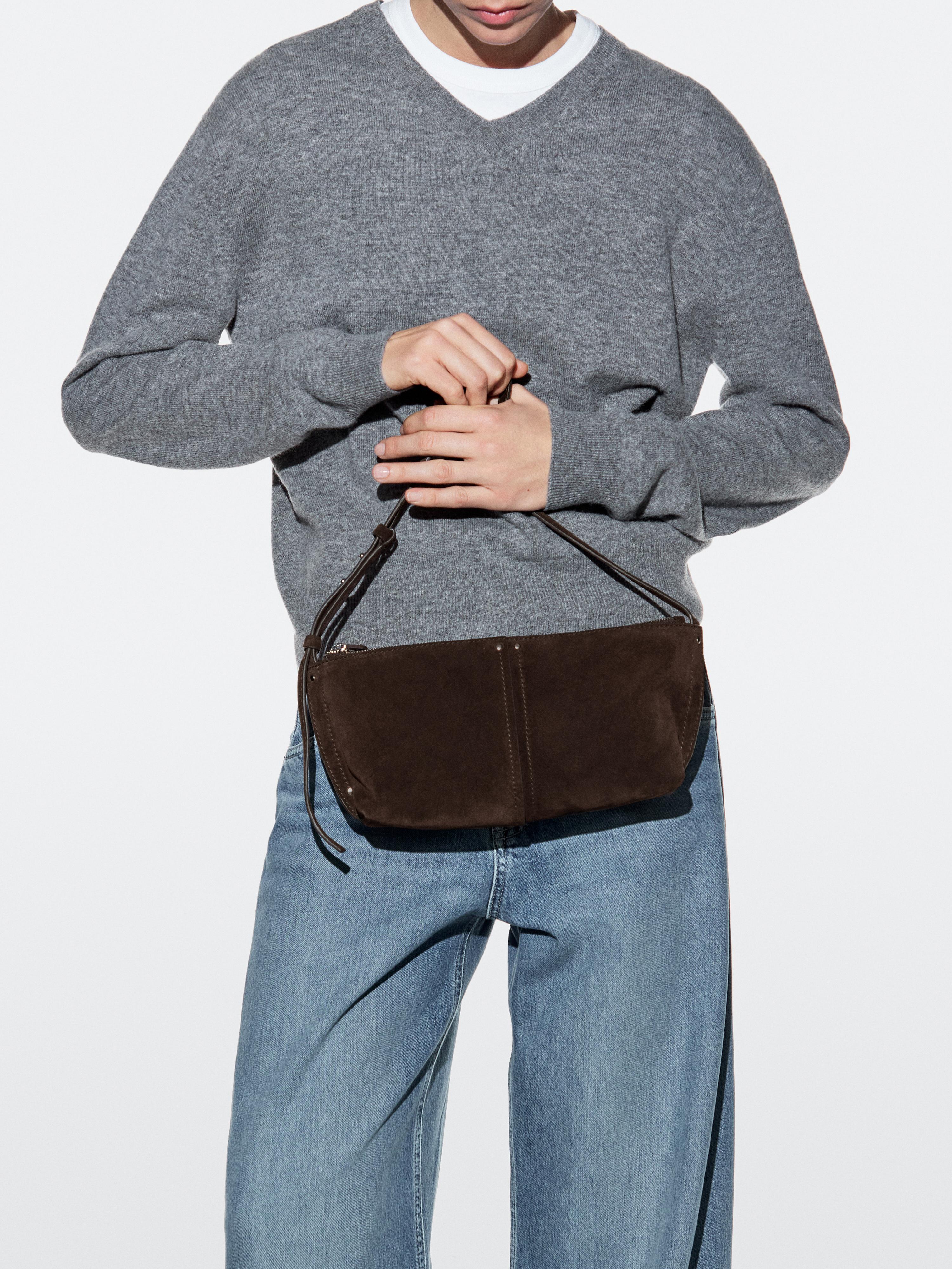 Plano medio de bolso marrón con cierre de cremallera y asa corta, combinado con suéter gris y jeans azules.