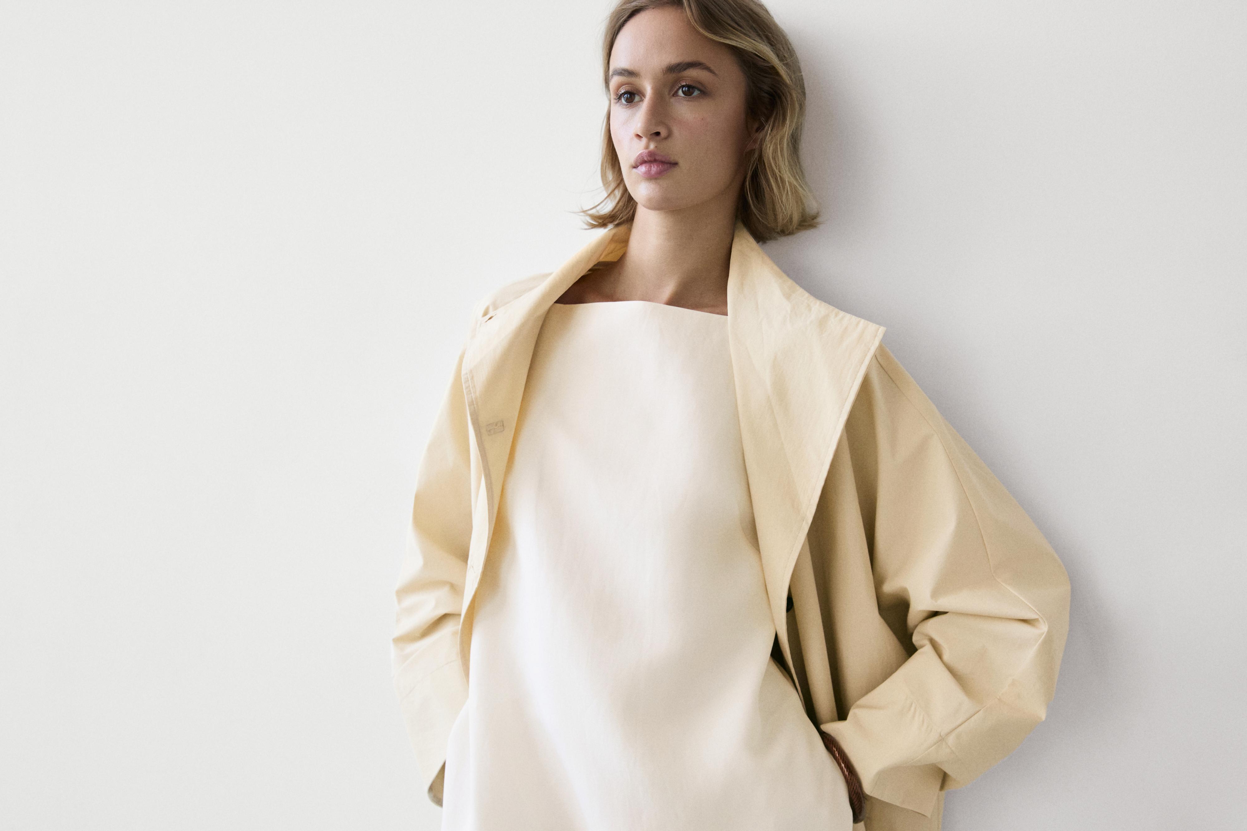 Plano medio de vestido mini branco sen mangas con escote cadrado, combinado con chaqueta beixe de colo amplo.