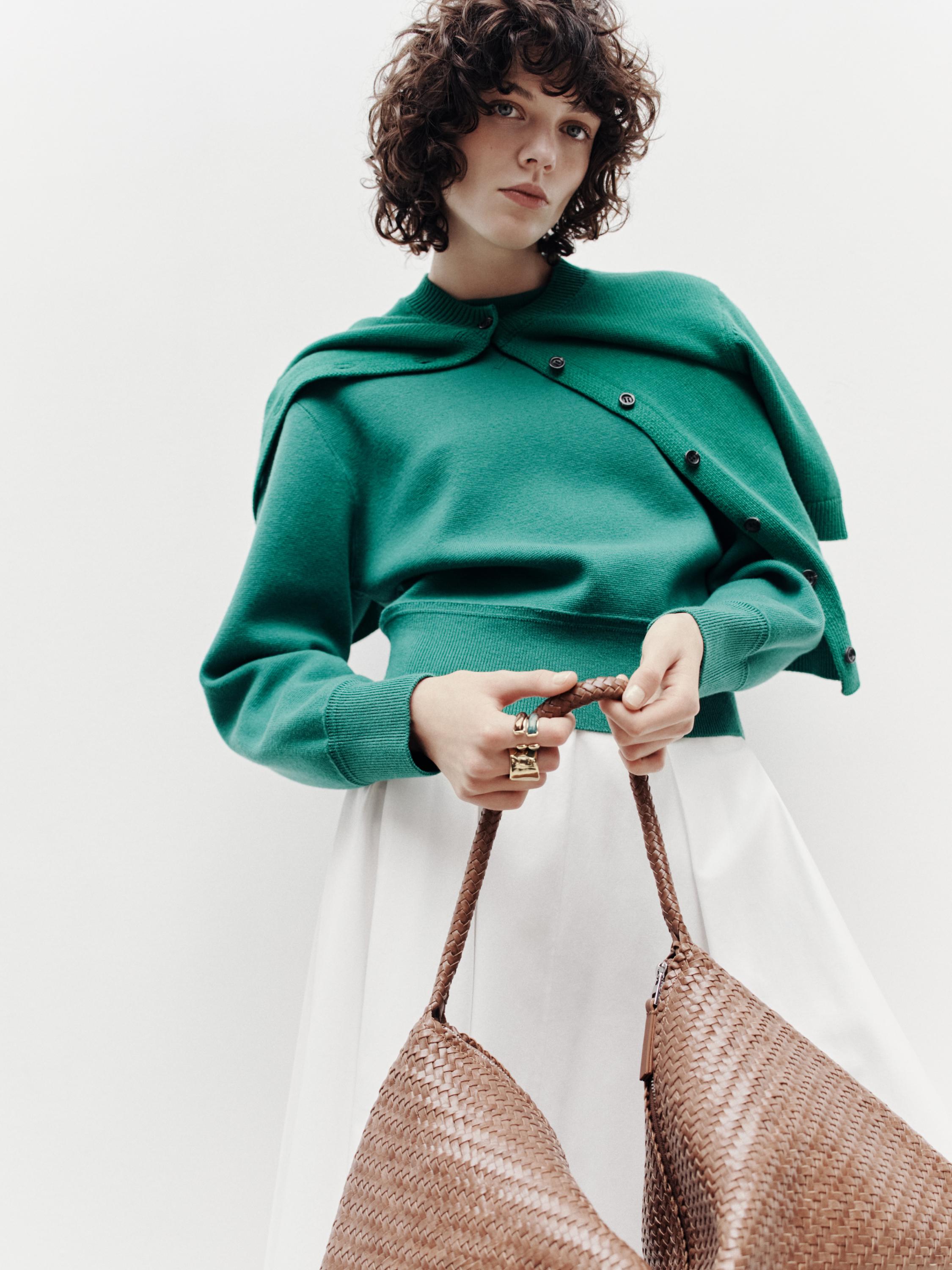 Средний план зеленого свитера с черными пуговицами и белой юбки. В руке плетеная коричневая сумка.