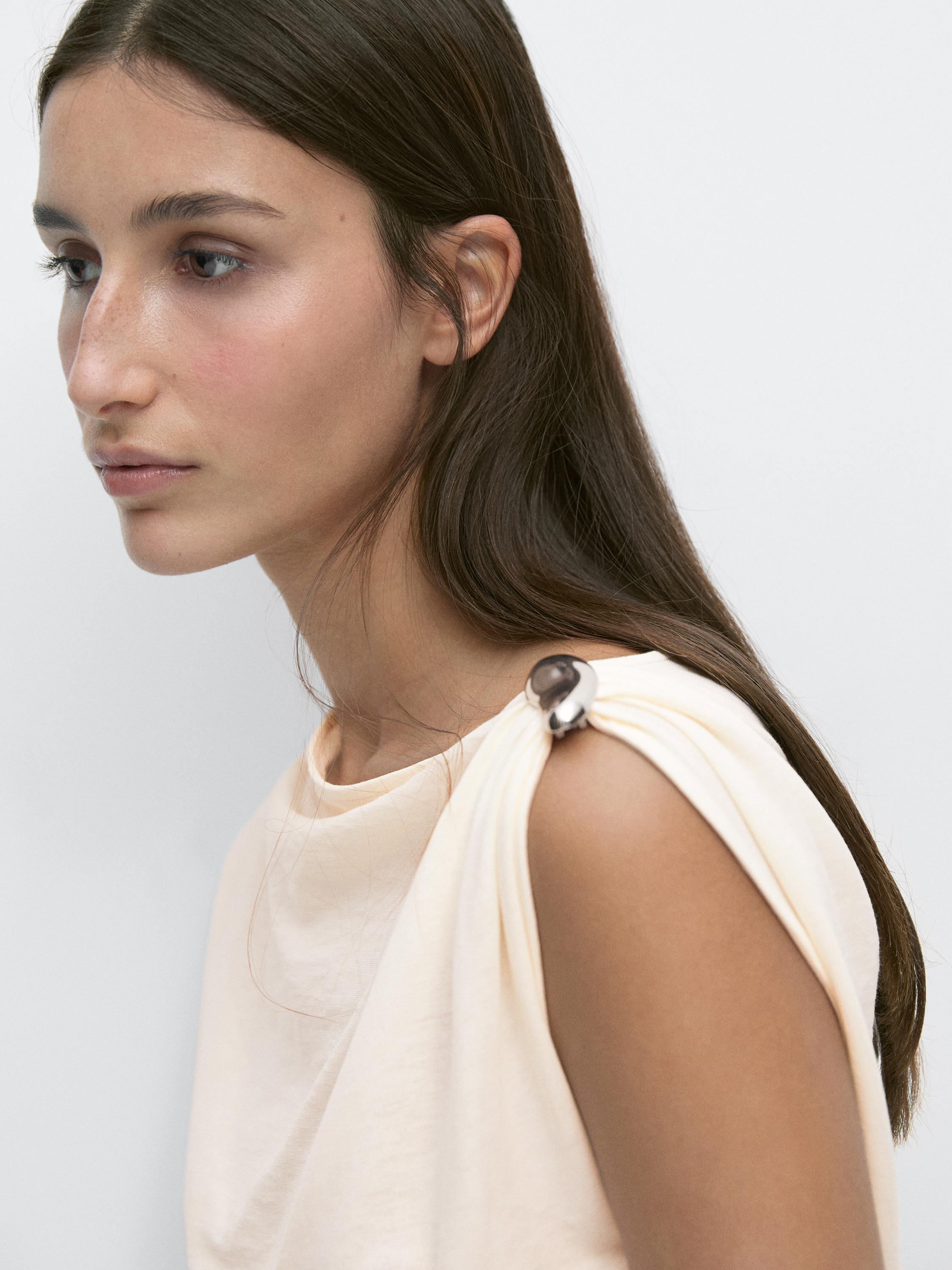 "Detalle de hombro de top blanco asimétrico con aplique metálico."