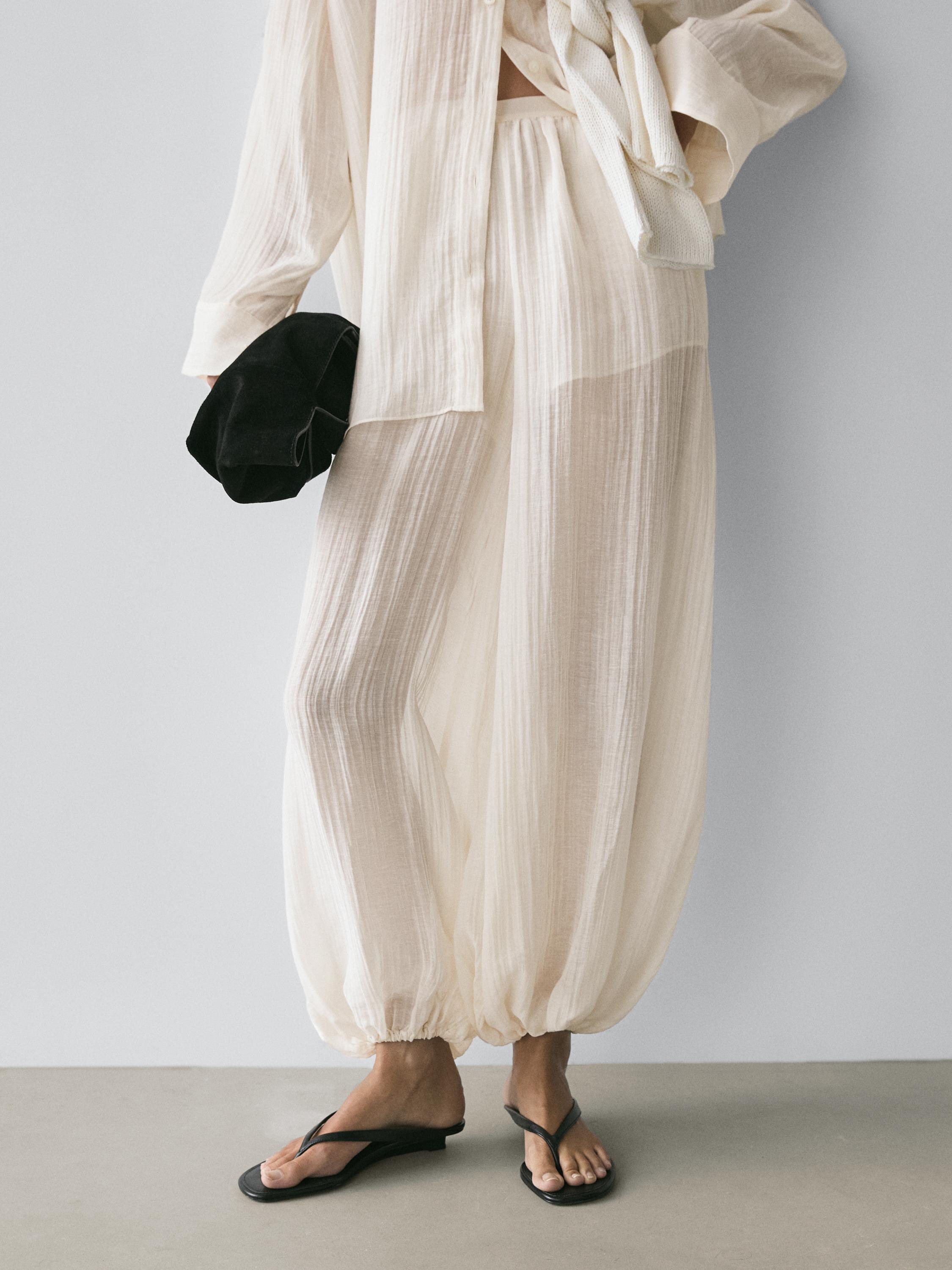 Pliisitud elevandiluuvärvi pluusi ja pükste komplekt, elastse vöökoha ja kitsaste säärtega püksid, mustad sandaalid.