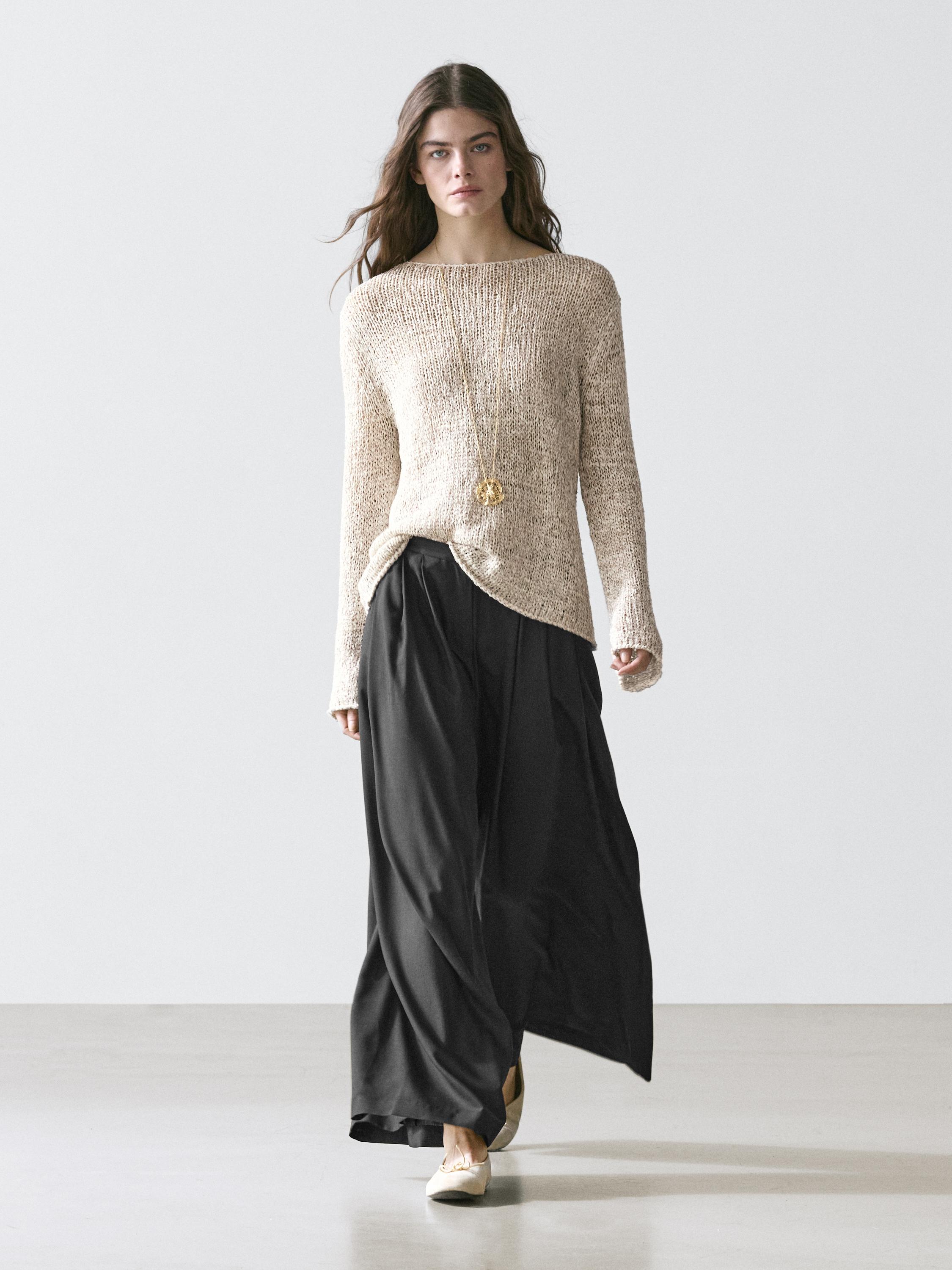 Conjunto de suéter beige de punto y pantalón negro fluido con pinzas, acompañado de colgante largo dorado.