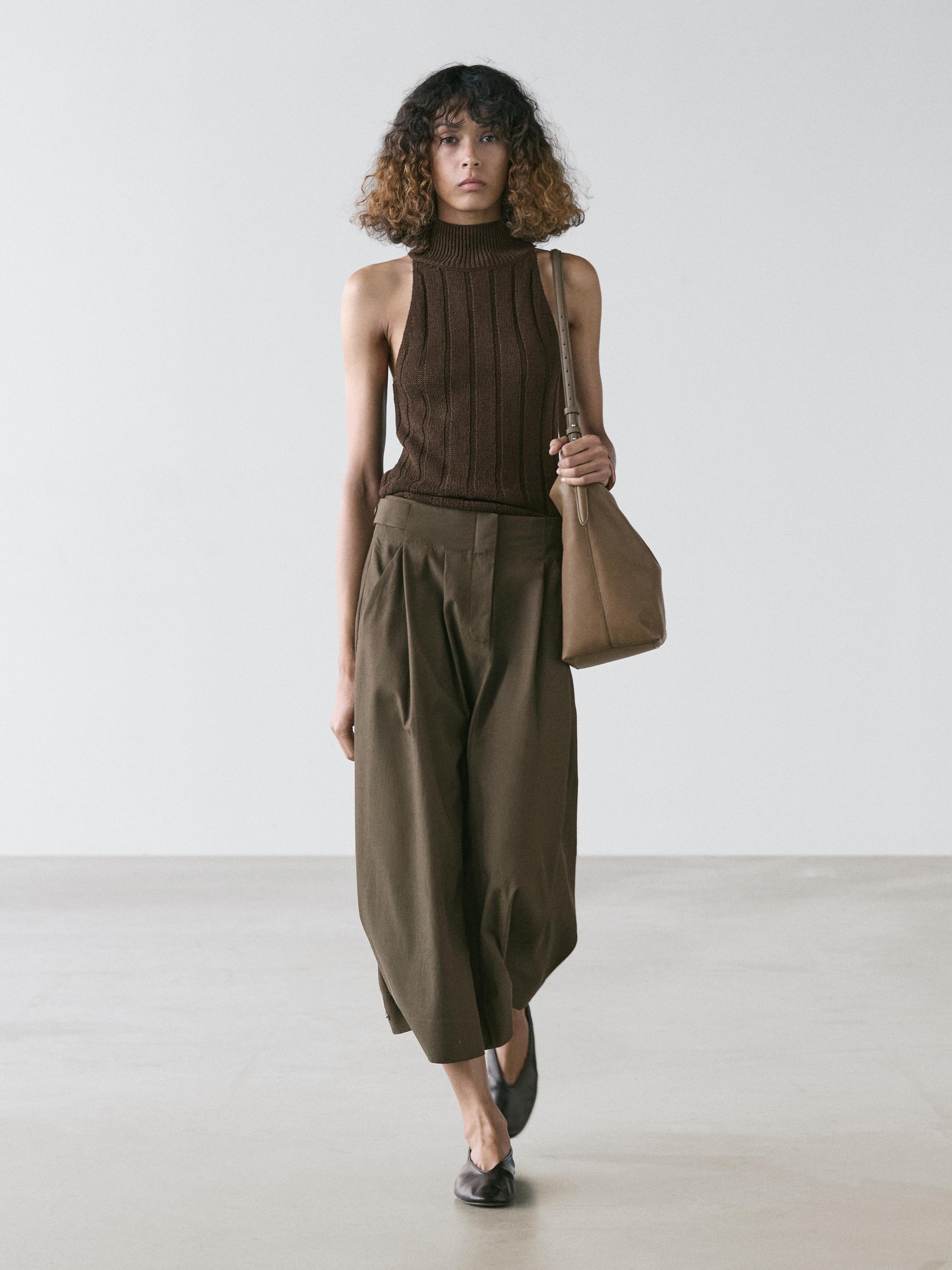 Mahoi marroi mahukarik gabeko eta kaki koloreko gona midi tolesduna, sorbaldako poltsa marroia eta zapata beltzak.