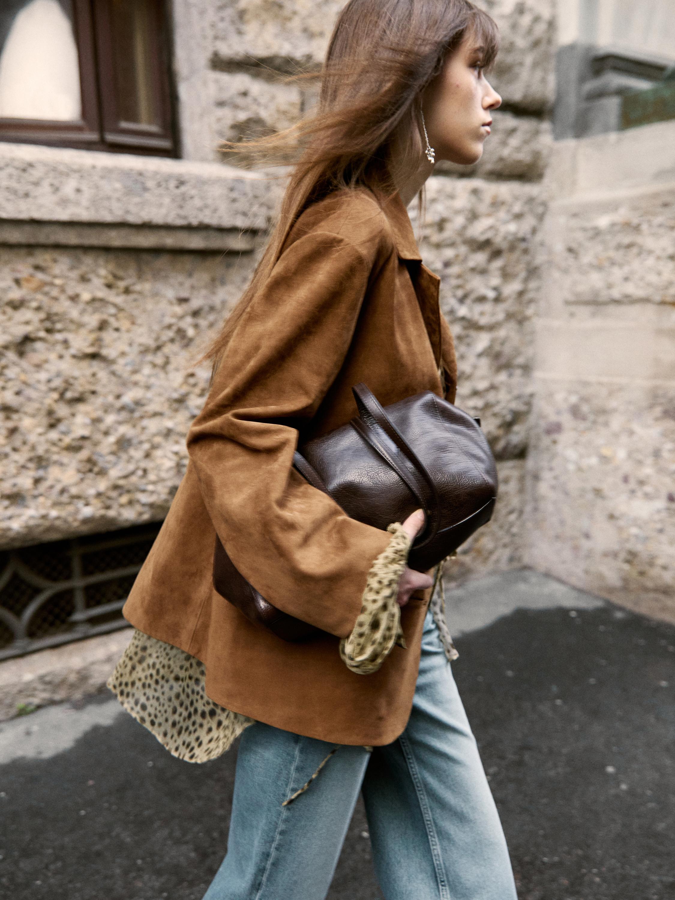Πλάνο μέσο από καφέ σακάκι με εμπριμέ μπλούζα ορατή στα μανίκια και μπλε τζιν. Σκούρα καφέ τσάντα στο χέρι.
