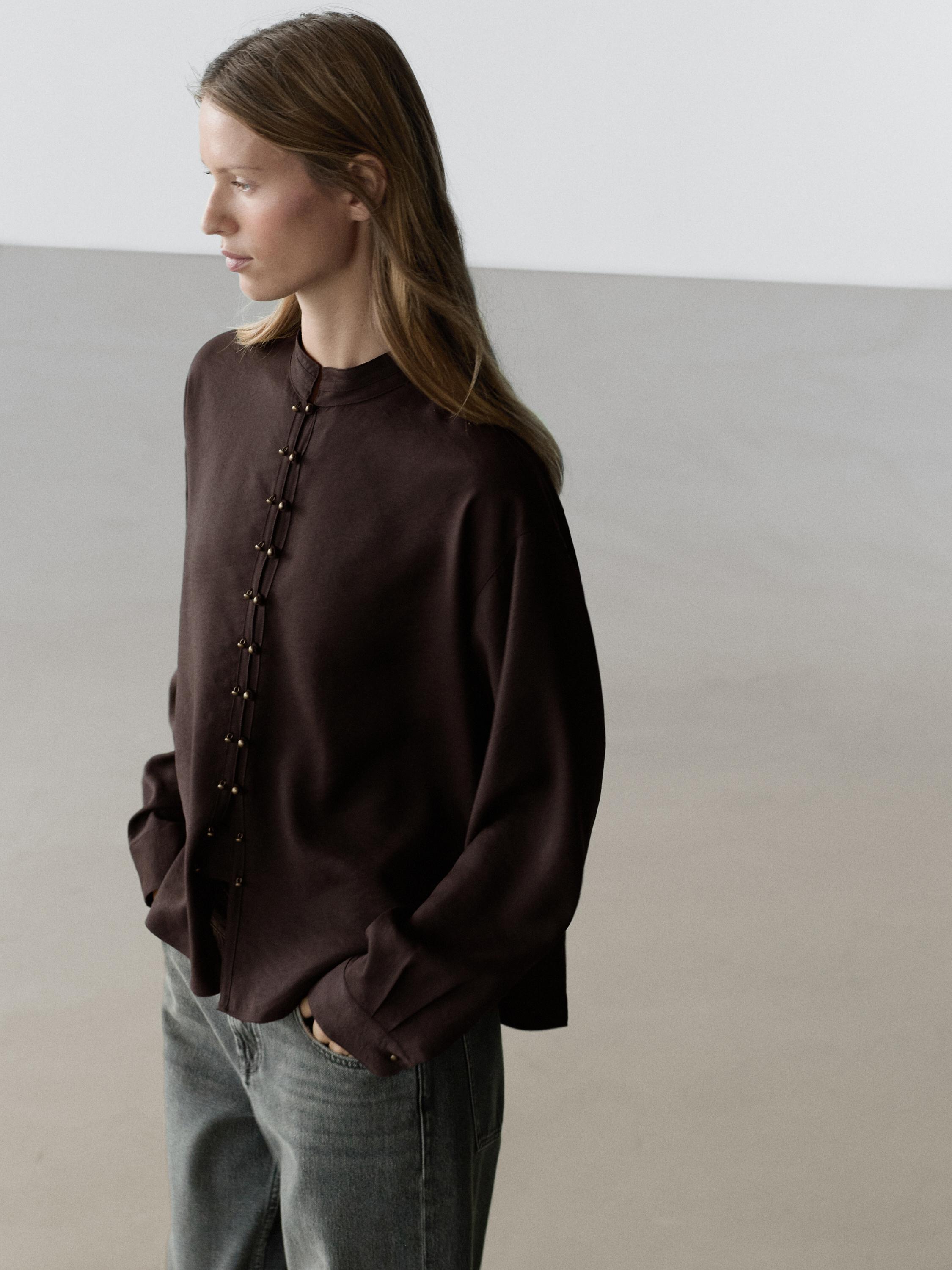 Mid-shot of a flowing terracotta shirt with front button detail and a mandarin collar, paired with gray pants.